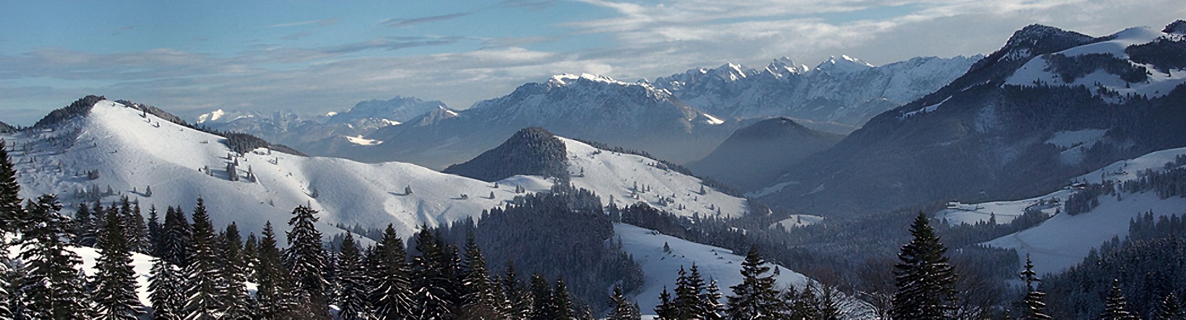 Panorama: verschneite Almwiesen und Fichtenwald, dahinter mehrere tief verschneite Bergketten unter blauem Himmel