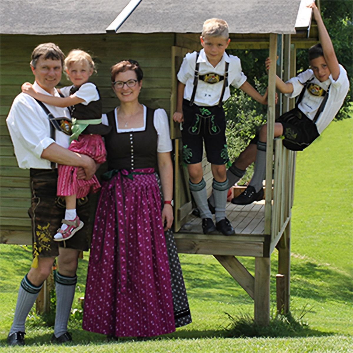 Familie Kellerer in Tracht vor einem hölzernen Spielhaus auf der Wiese: Alfons mit dem jüngsten Kind auf dem Arm, daneben Marlene im Dirndl, zwei Buben in Lederhosen stehen und hängen am Spielhaus