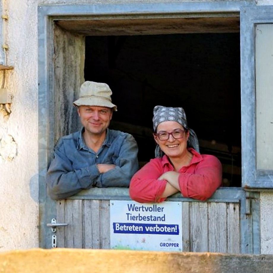 Alfons in a hat and Marlene in a headscarf and glasses leaning out of the open cowshed door and smiling, a sign reading “Wertvoller Tierbestand” (valuable livestock) on the door in front of them