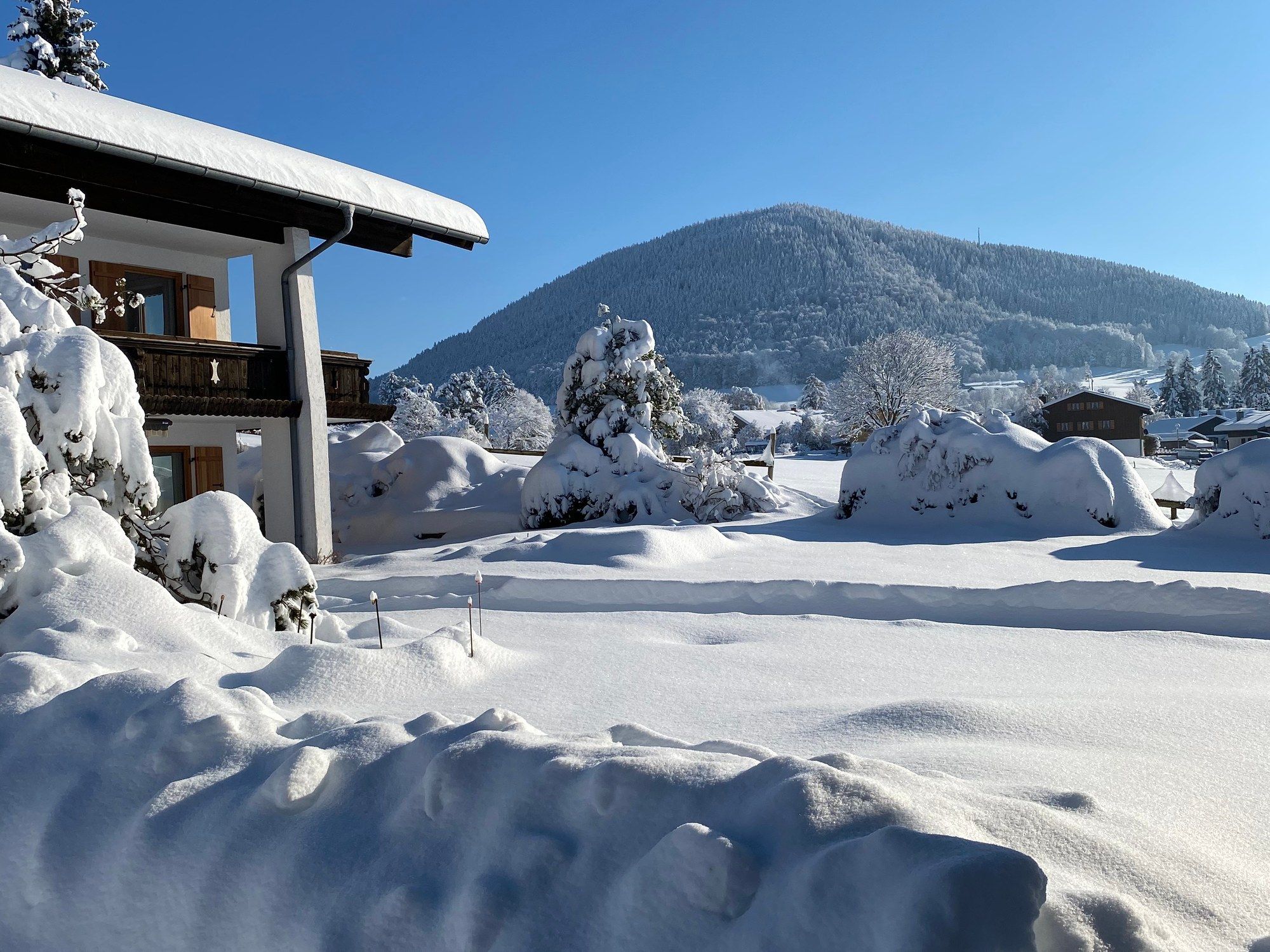 Blick vom Balkon im Winter über den tief verschneiten Garten auf einen bewaldeten Berg, links die Hausecke mit Schnee auf dem Dach