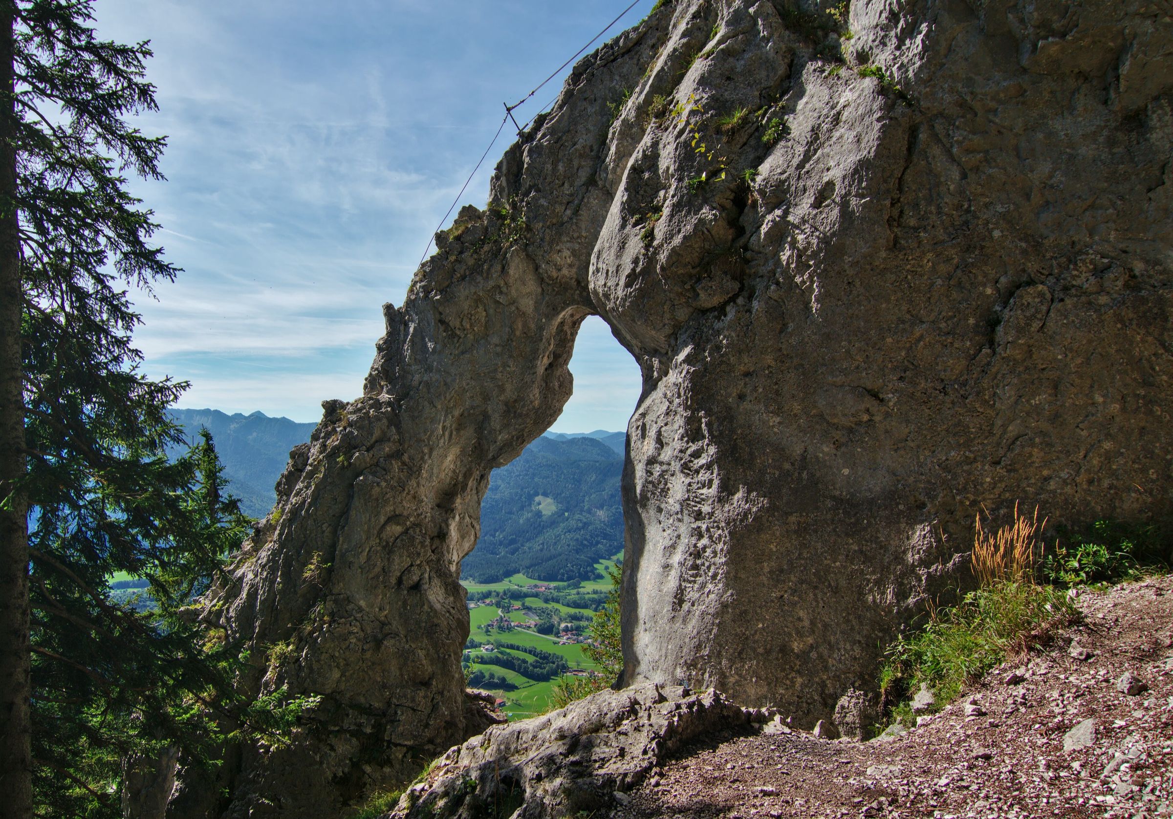 Ein großes Felstor, durch das man ins Tal hinunterschaut, davor steiniger Boden und Fichten