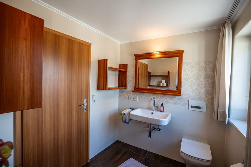 Bad mit Waschbecken unter einem holzgerahmten Spiegel, Wandregal, wandhängendem WC und Fliesen mit Ornamentmuster