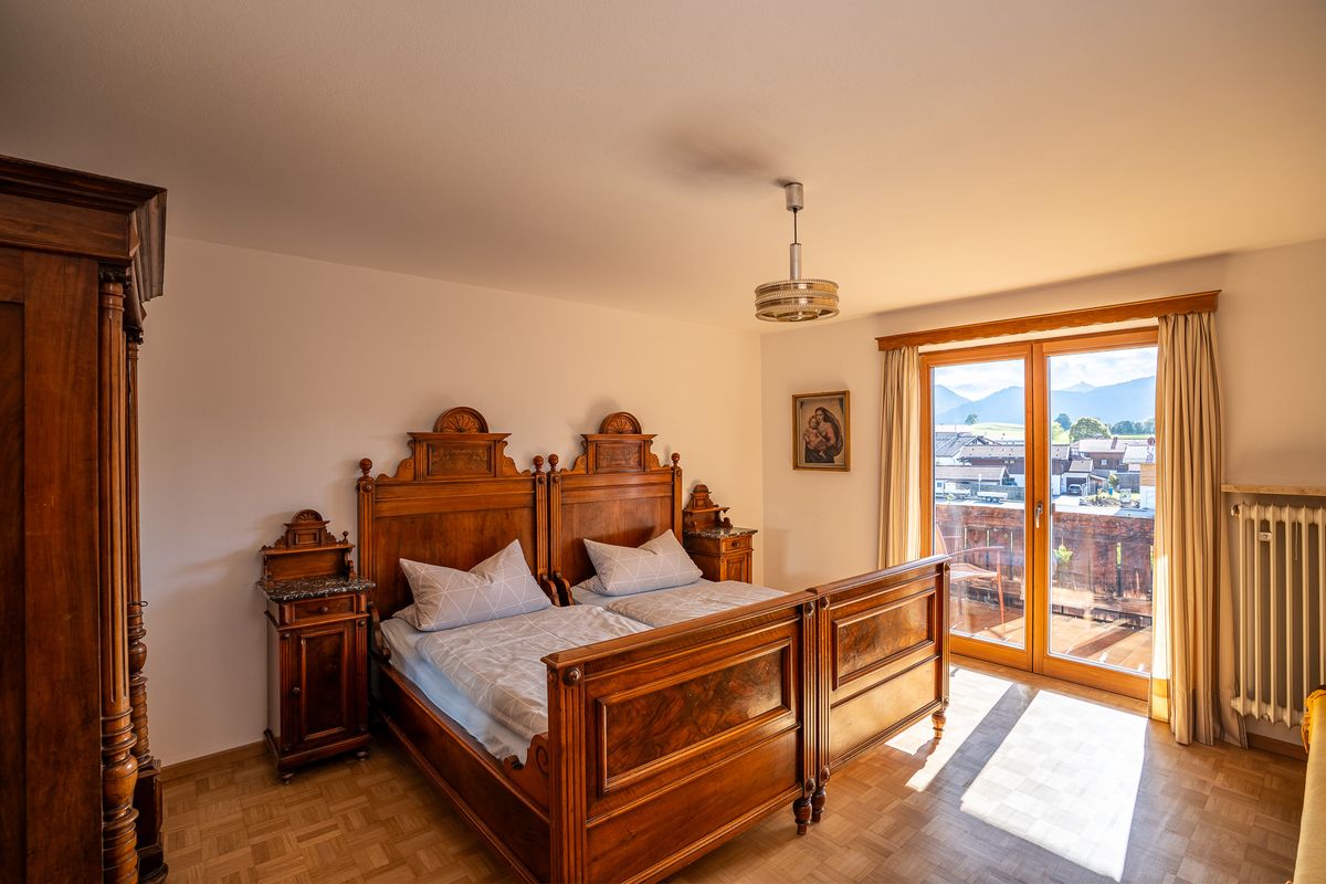 Schlafzimmer mit Doppelbett aus der Gründerzeit mit geschnitzten Kopfteilen und Nachtkästchen mit Marmorplatte, rechts die Balkontür mit Blick über die Dächer auf die Berge
