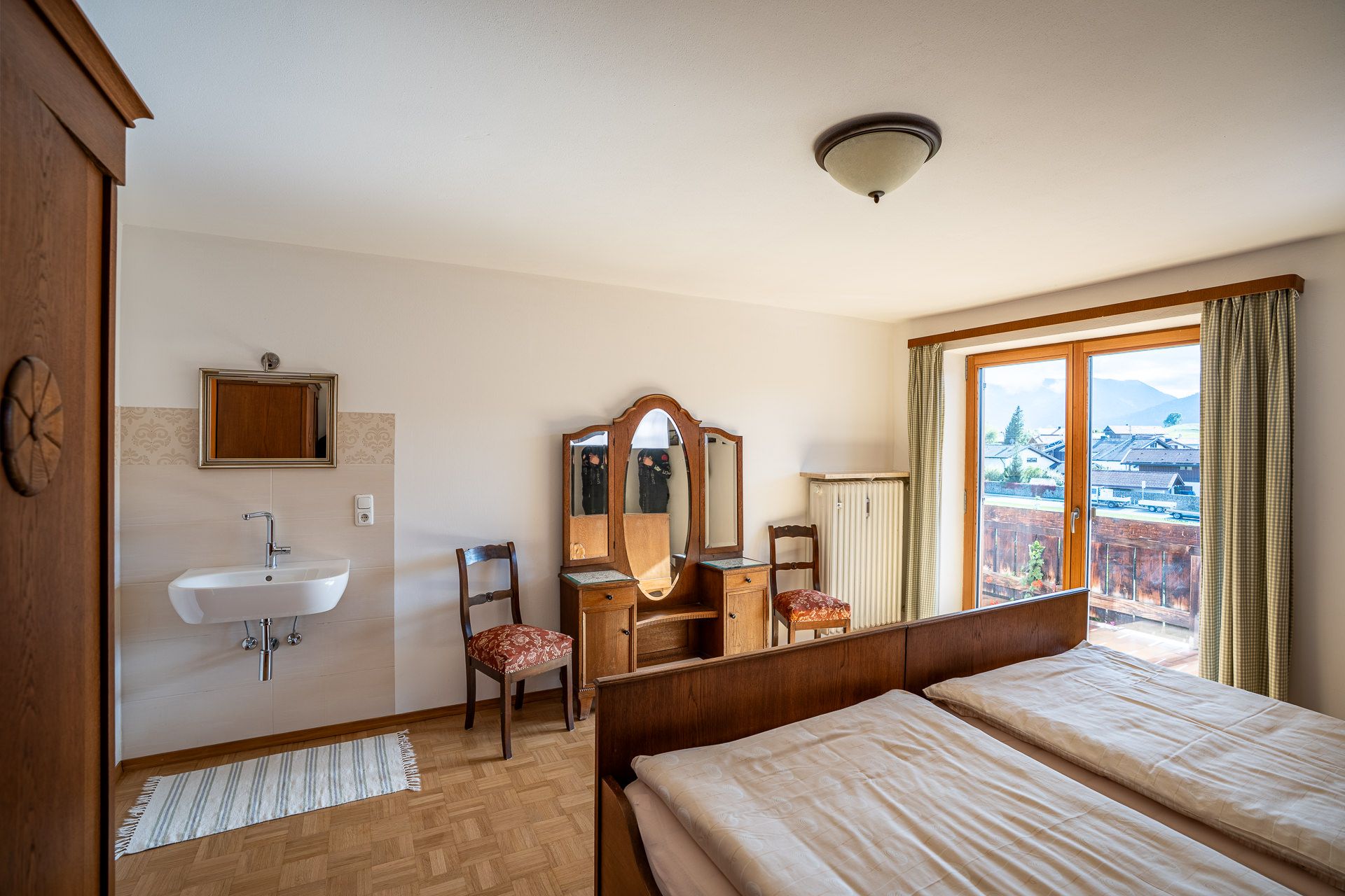 Zweites Schlafzimmer mit Frisiertisch mit dreiteiligem Spiegel, zwei gepolsterten Stühlen und einem Waschbecken, rechts die Balkontür mit Blick über Dächer auf die Berge