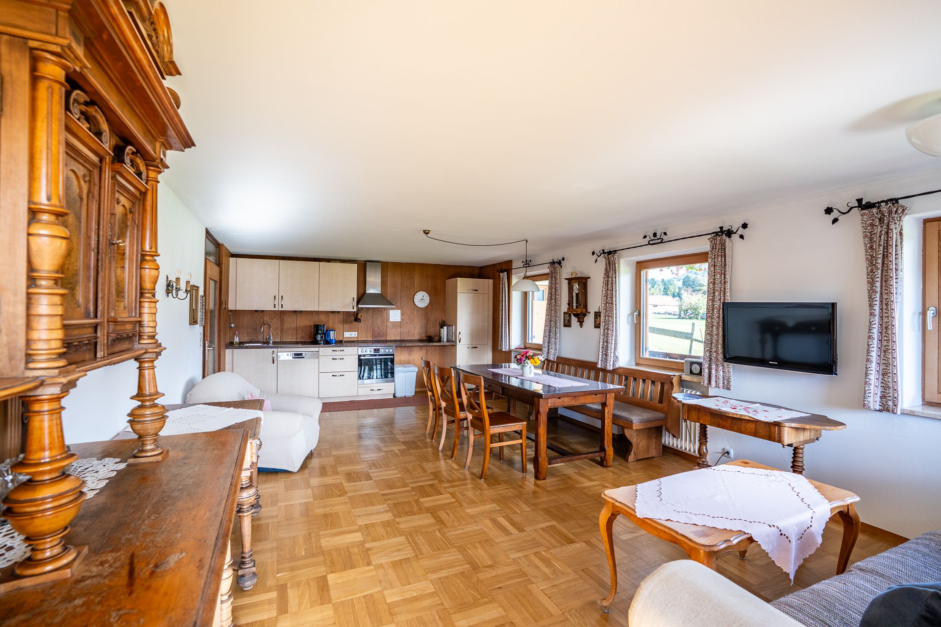 Die ganze Wohnküche mit Küchenzeile, Backofen und Dunstabzug hinten, davor der lange Esstisch mit Bank, rechts der Fernseher, vorn links ein geschnitzter antiker Schrank