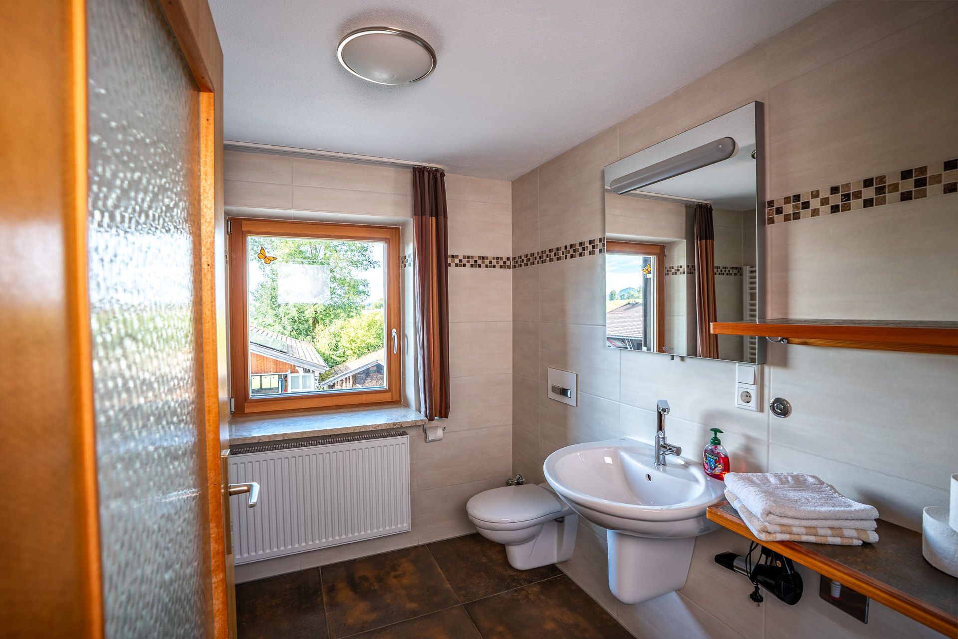 Bathroom with a window, a radiator, a wall-hung WC, a washbasin below a large mirror and a wooden shelf with towels and a hairdryer