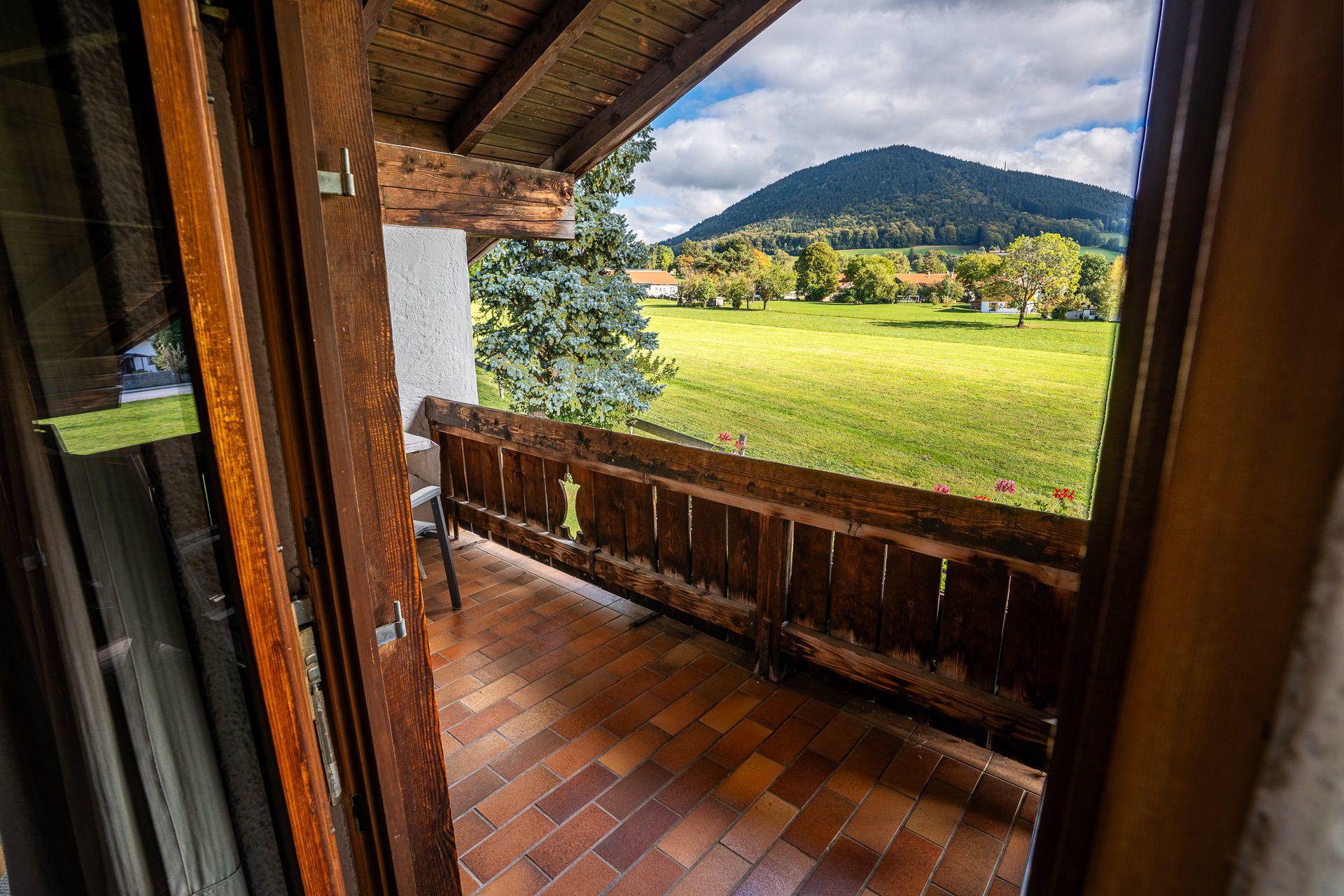 Balkon mit Holzgeländer und Fliesenboden, Blick über eine grüne Wiese auf einen bewaldeten Berg, dazwischen Bäume und Häuser