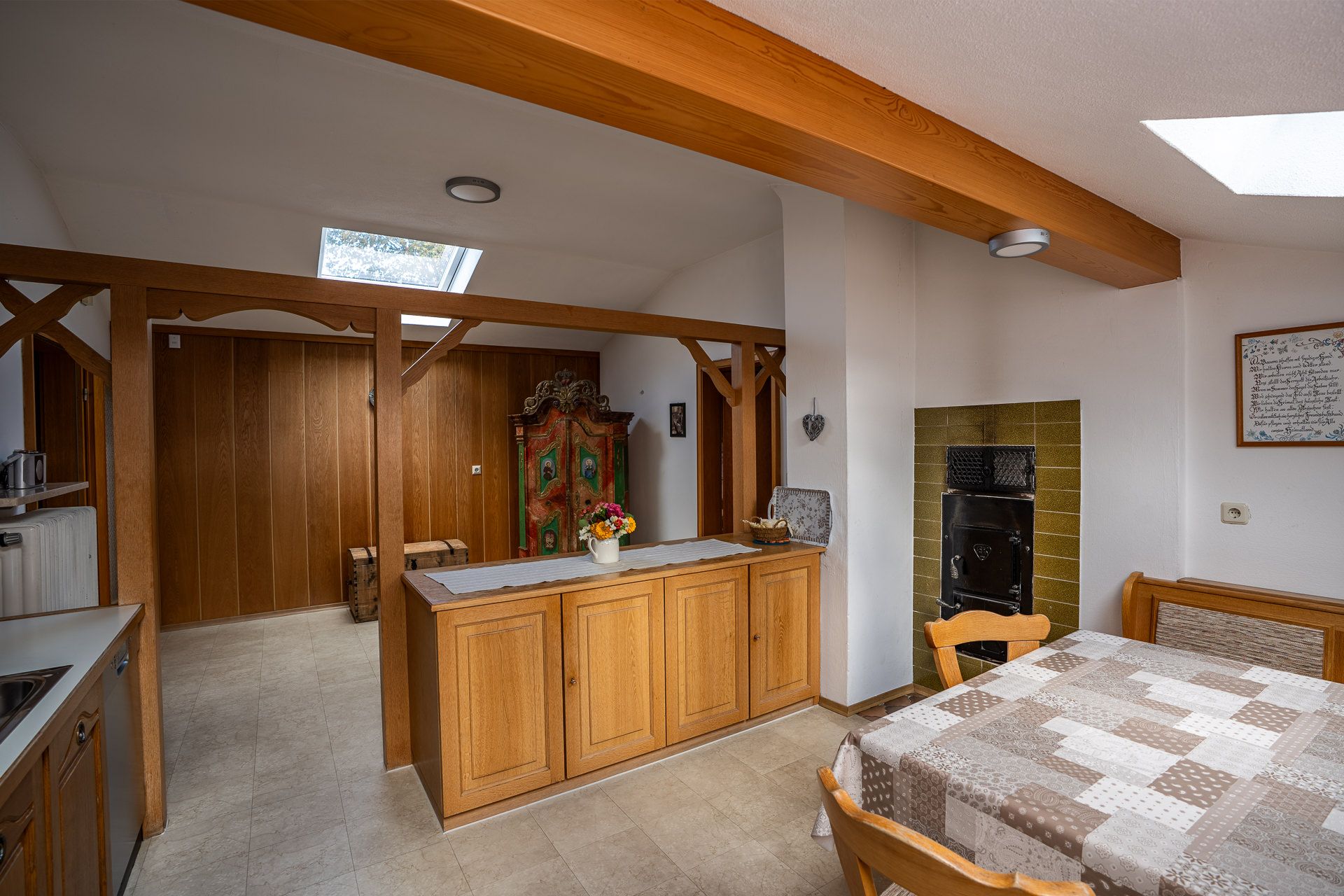 Kitchen with a wooden room divider and base cupboard, a wood-panelled wall and a painted farmhouse cupboard at the back, a green-tiled stove recess with stove doors and the dining table with a corner bench on the right, and light from the roof windows