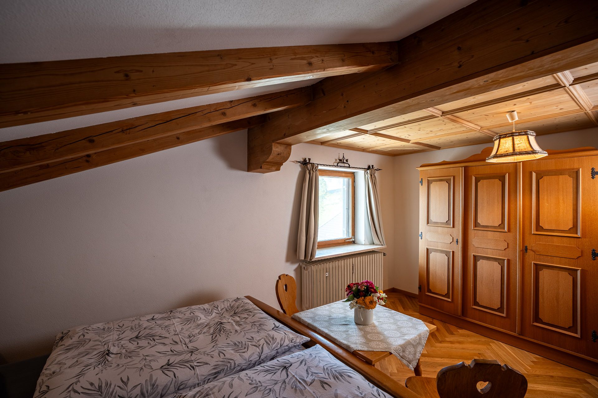 Farmhouse wardrobe under the wooden ceiling and its beams, a window with a radiator and the small table with flowers, the double bed in front