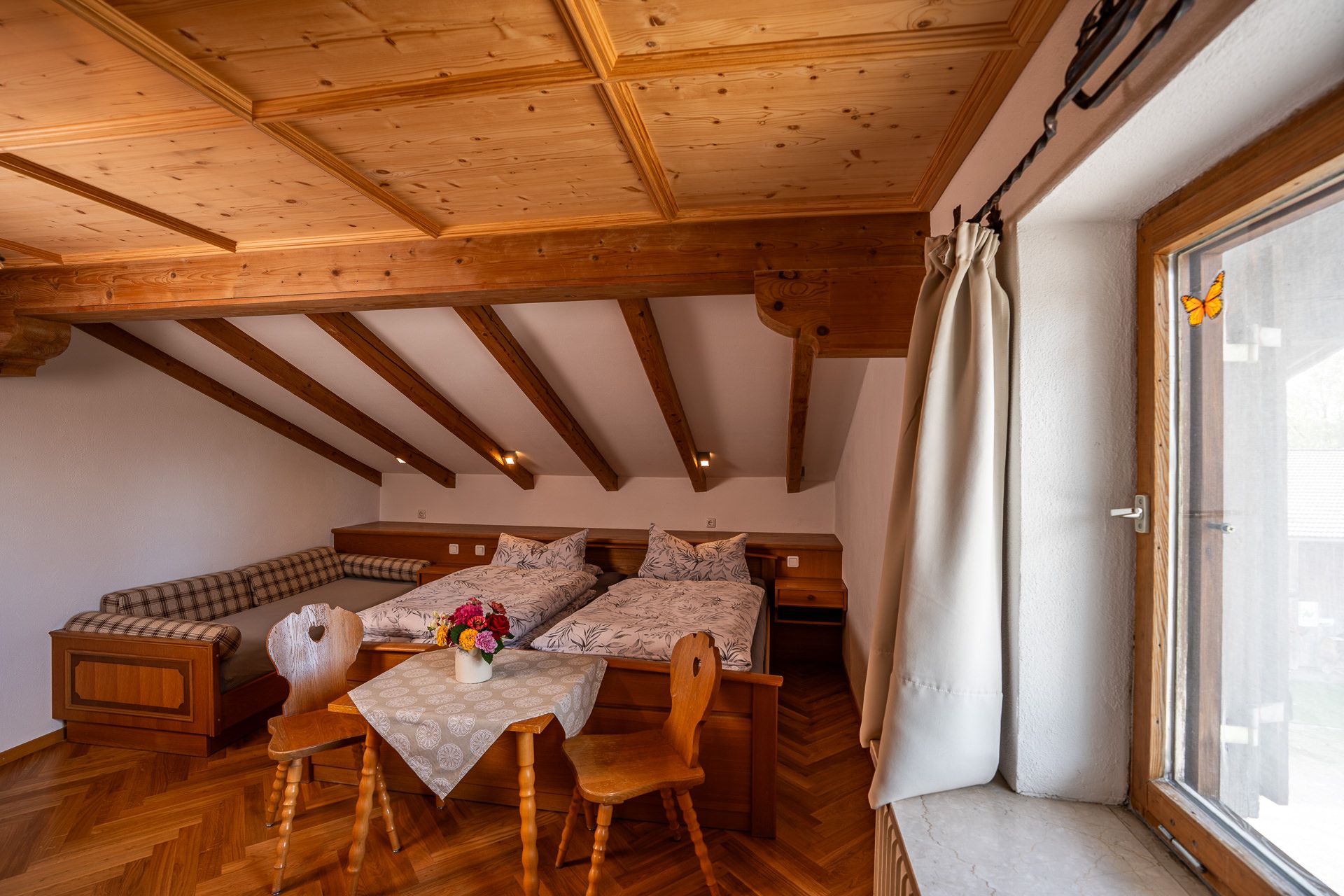 Bedroom with a coffered wooden ceiling and beams, a double bed, a daybed with checked cushions and a small table with two chairs by the window