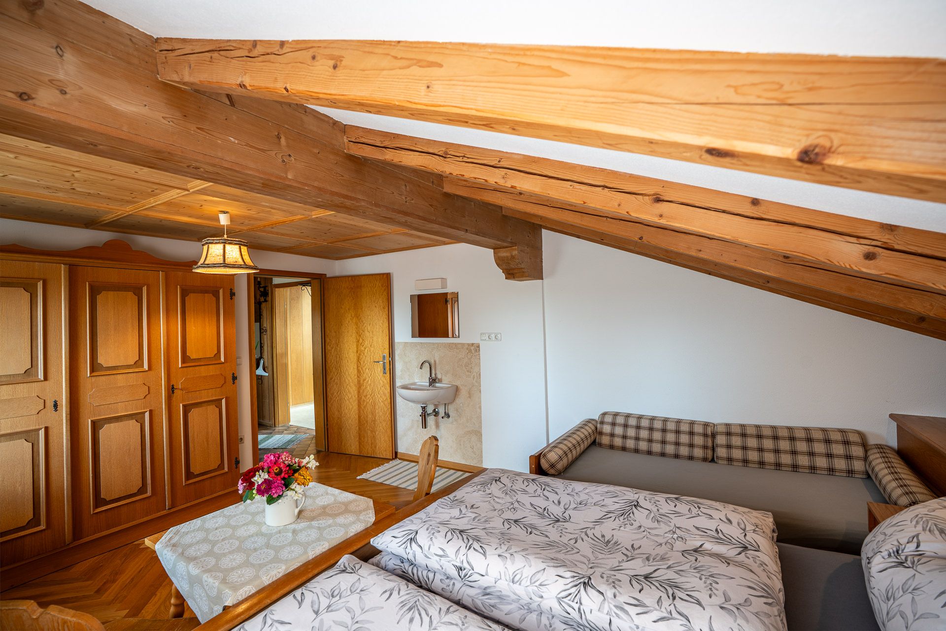 The same bedroom under exposed beams with a farmhouse wardrobe, a washbasin and mirror beside the door, and the double bed and the daybed with its checked cushions in front