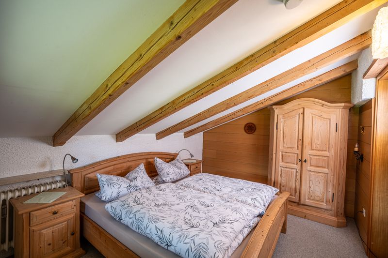 Second bedroom under the sloping roof with exposed beams, a light wooden double bed, bedside cabinets with reading lamps and a farmhouse wardrobe against the wood-panelled wall