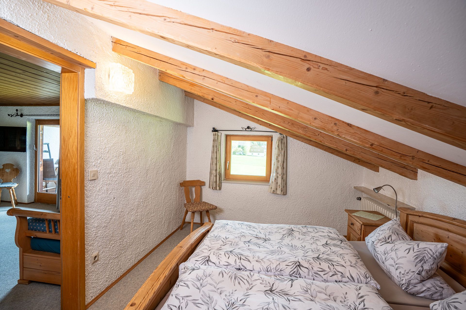 Second bedroom with beams under the sloping roof, a small window looking out onto greenery, a chair and the double bed, the open door to the parlour on the left