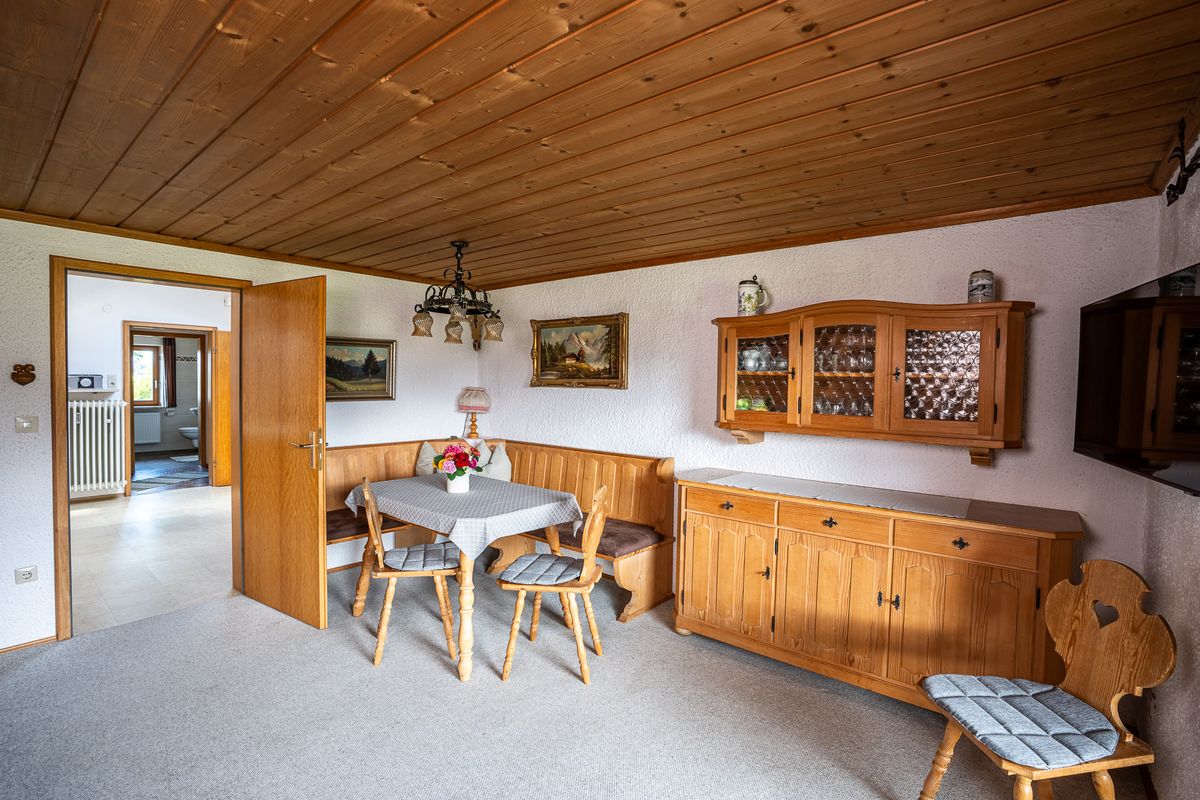 Wohnstube mit Holzdecke, Eckbank und Tisch, einer Anrichte und einem Wandschrank mit Glastüren, rechts ein Stuhl mit Herz in der Lehne
