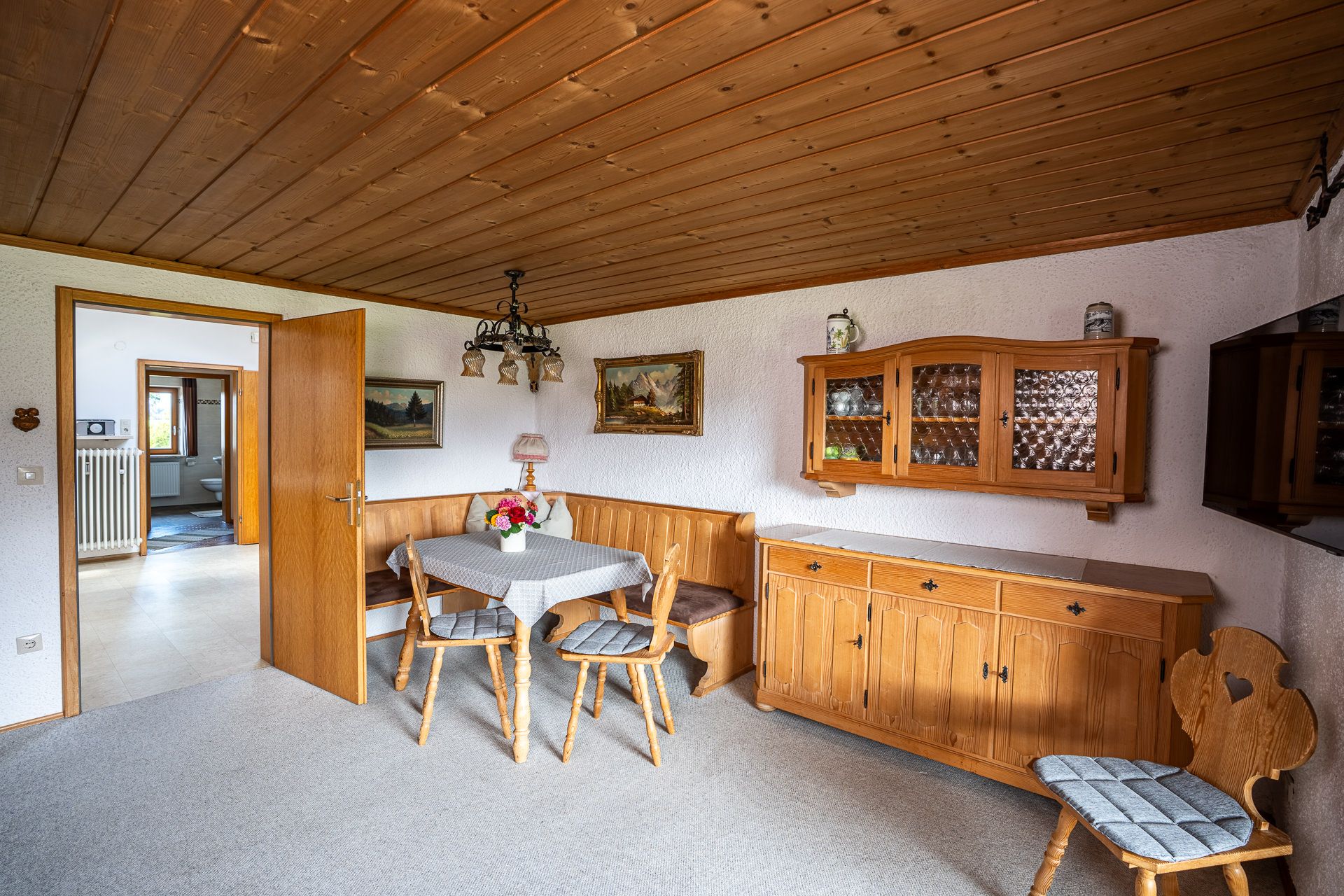 Parlour with a wooden ceiling, a corner bench and table, a sideboard and a glass-fronted wall cabinet, and a chair with a heart cut into the back on the right