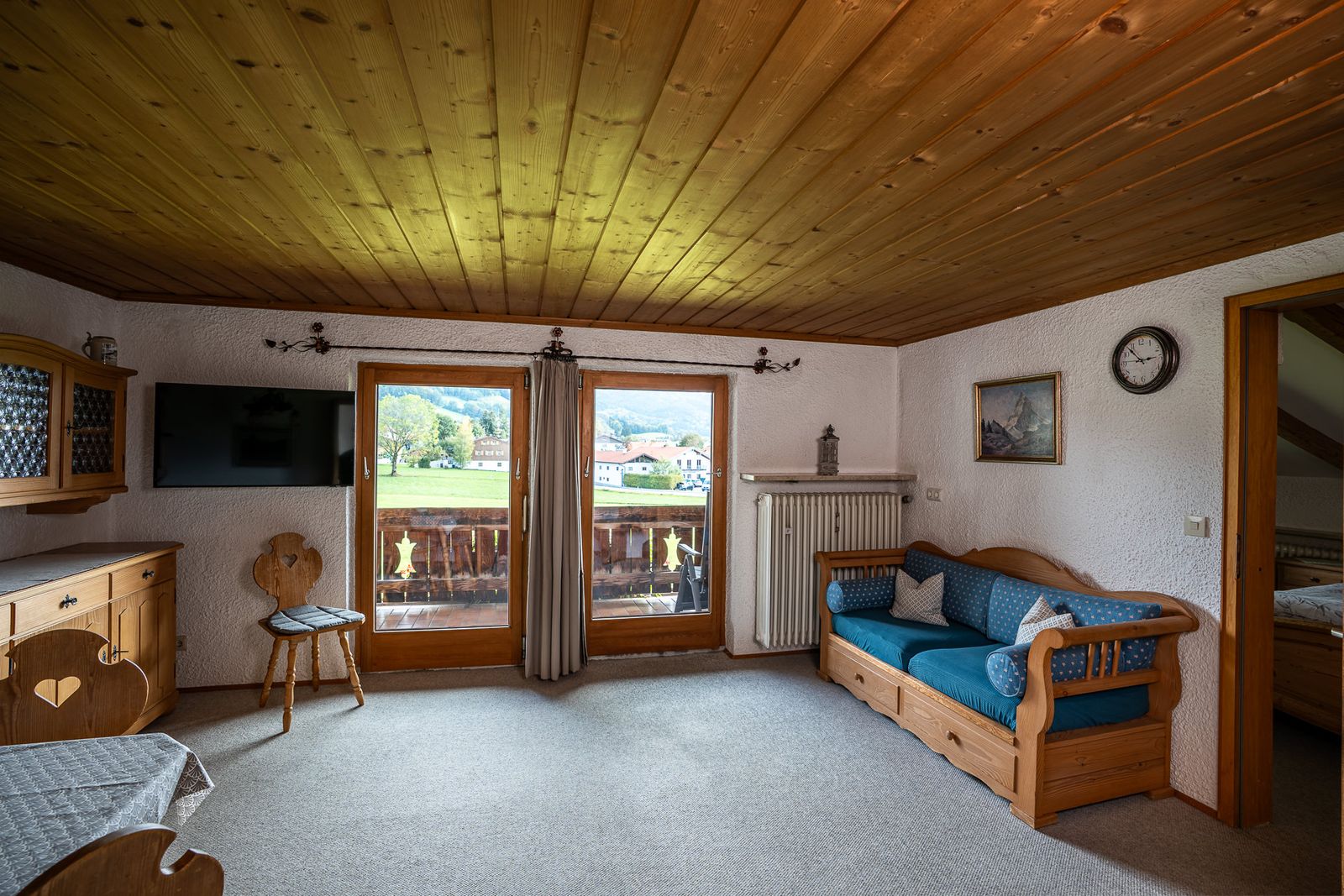 Parlour of the top-floor apartment with a wooden ceiling and a blue sofa, with a view through the balcony door across the meadow