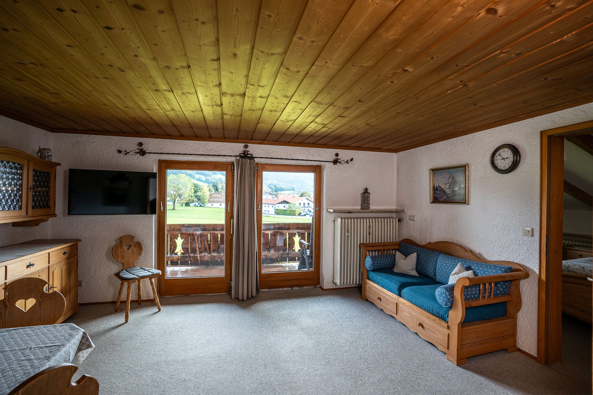 Wohnstube der Ferienwohnung im Dachgeschoss mit Holzdecke, blauem Sofa und Fernseher, durch die Balkontür sieht man über die Wiese zu Häusern und Bäumen