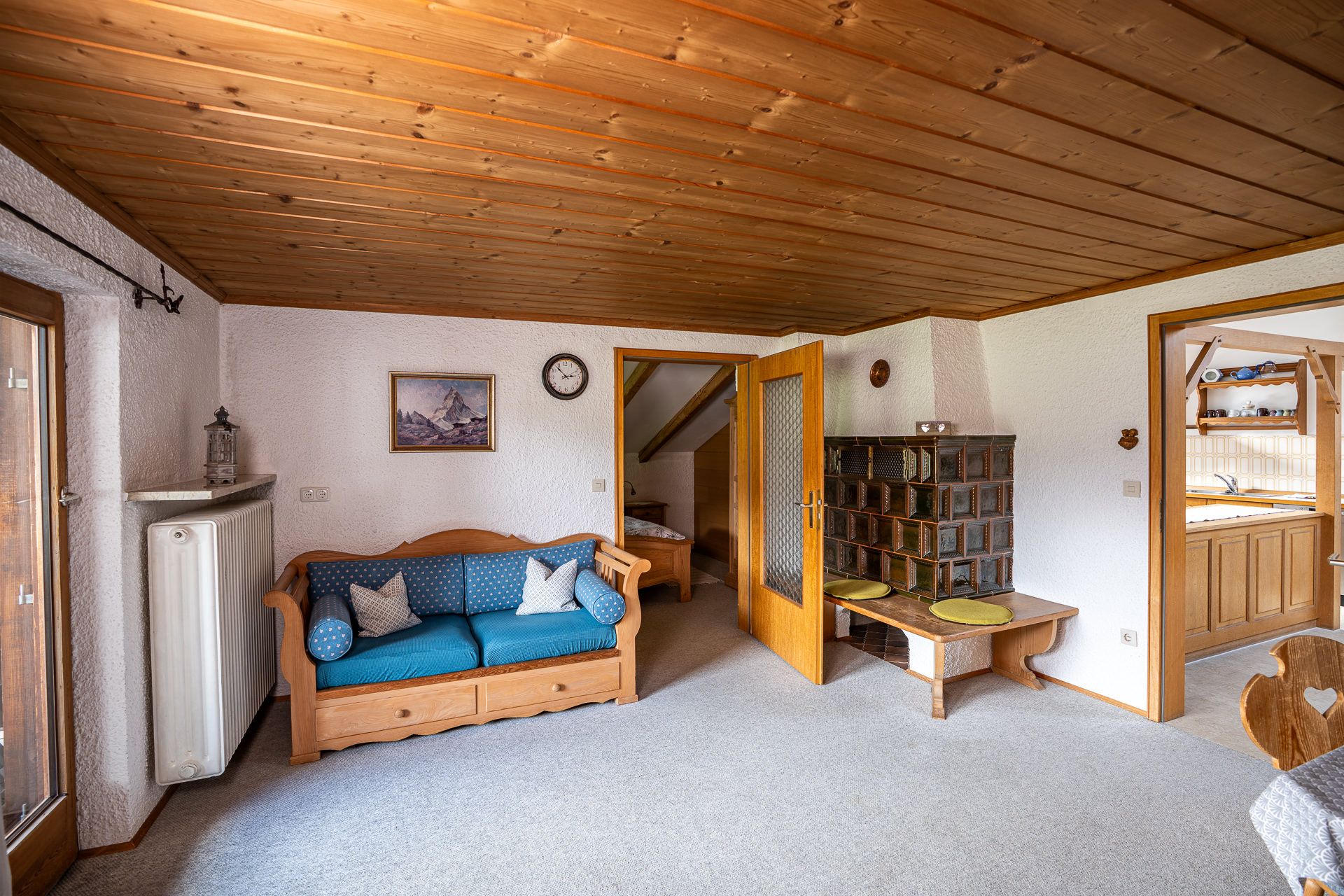Blue sofa in the parlour under the wooden ceiling, with a brown tiled stove and bench on the right