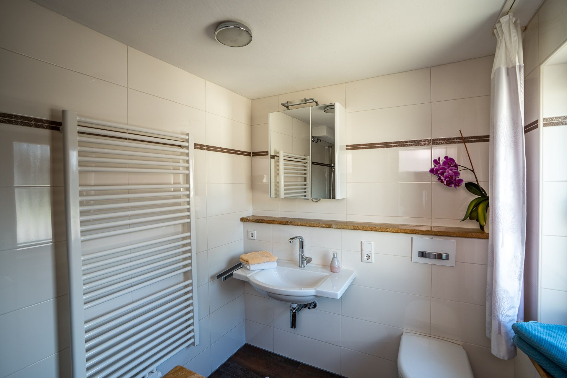 Bad mit Waschbecken unter einem Spiegelschrank, wandhängendem WC, Handtuchheizkörper und einer Orchidee auf der Ablage
