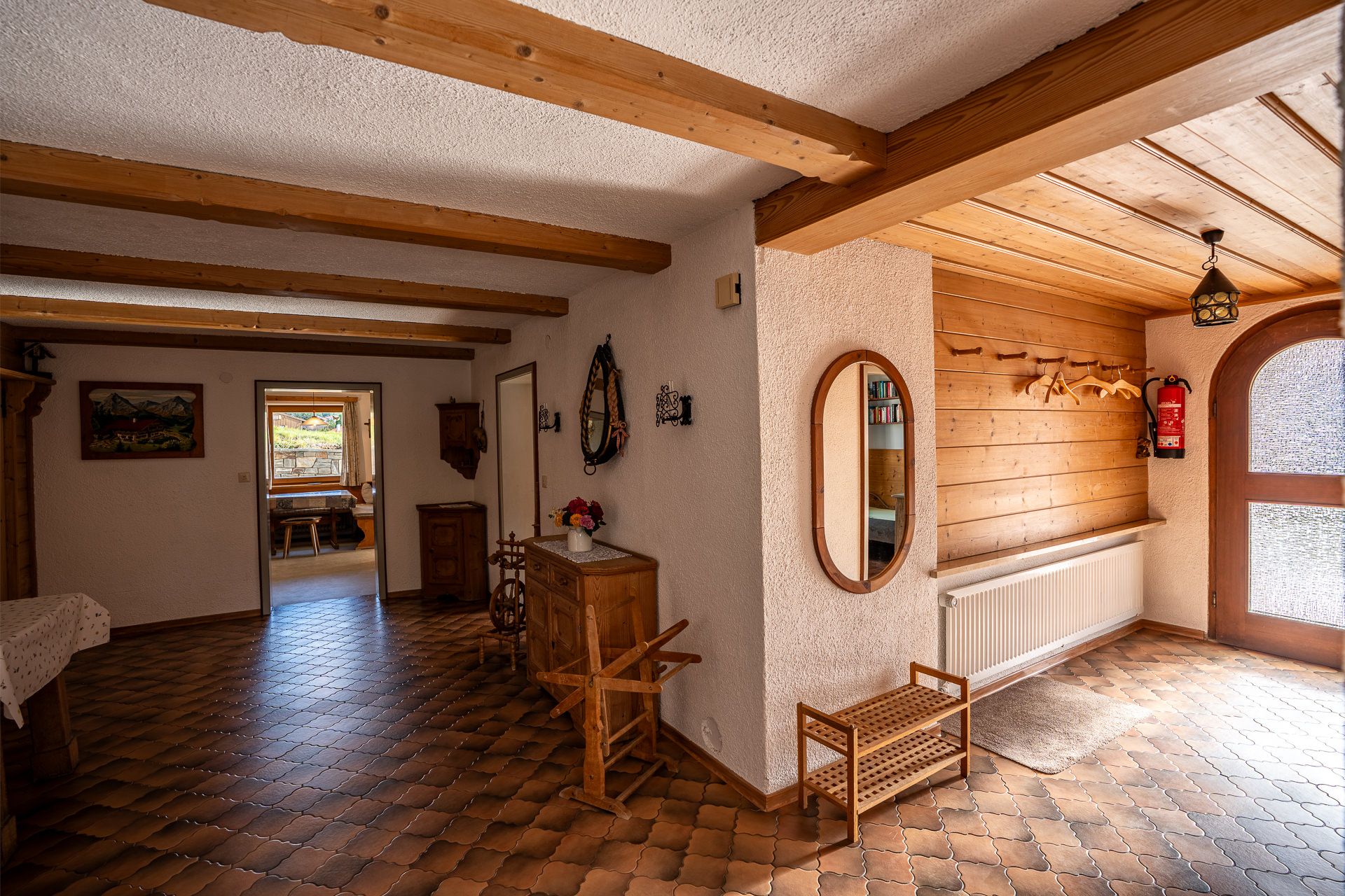 Diele mit Holzbalken und Fliesenboden, an der Holzwand Garderobenhaken, Heizkörper und ein Schuhregal, rechts die Eingangstür mit Rundbogen, hinten der Blick in die Küche