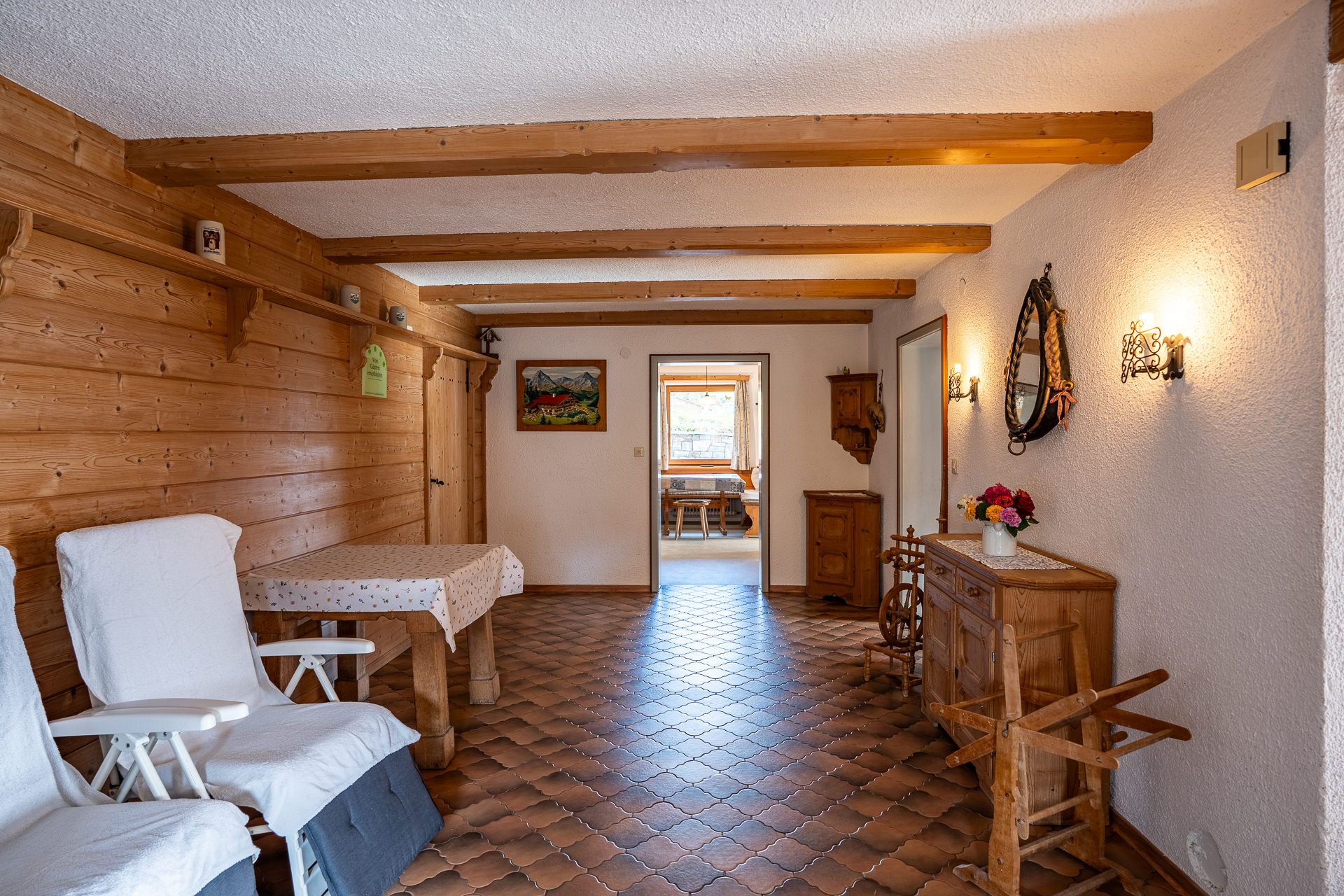 Diele mit Holzbalken, an der Holzwand ein Tisch und zwei Gartenstühle mit Auflagen, rechts ein Spinnrad und eine Kommode mit Blumen, hinten der Blick in die Küche