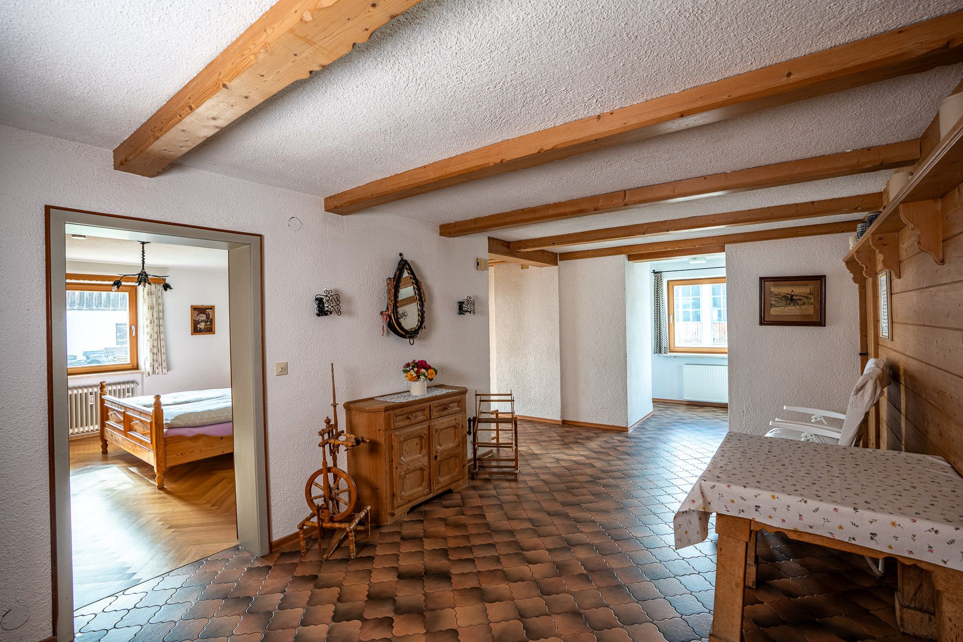 Diele mit Holzbalken und Fliesenboden, ein Spinnrad und eine Kommode mit Blumen, links durch die offene Tür das Schlafzimmer mit dem Holzbett, hinten ein Fenster