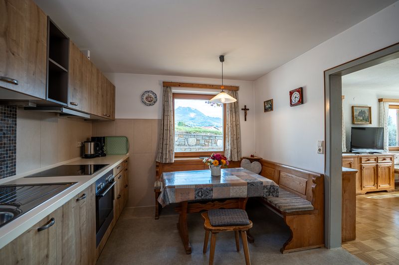 Küche in Holzoptik mit Kochfeld, Backofen und Hängeschränken, am Fenster die Essecke mit Eckbank, draußen eine Steinmauer und ein Berg, rechts der Blick ins Wohnzimmer