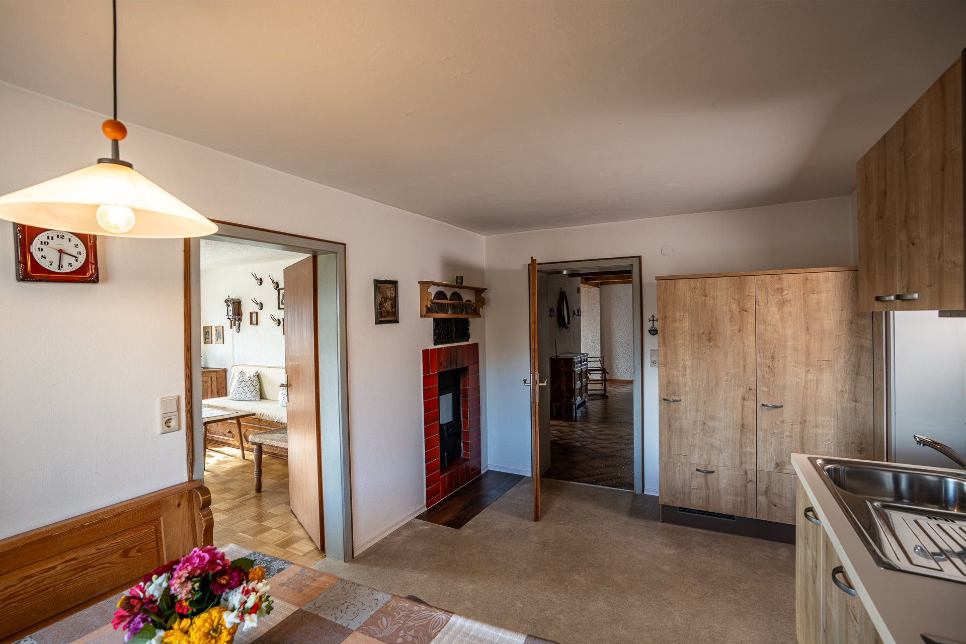 Küche mit Hängelampe über dem Esstisch, einer rot gekachelten Ofennische mit Ofentür, einem hohen Schrank in Holzoptik und der Spüle, durch die offenen Türen sieht man ins Wohnzimmer mit dem Sofa und in die Diele