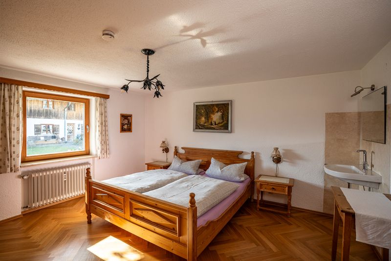 Schlafzimmer mit Doppelbett aus hellem Holz, zwei Nachttischen und Fischgrätparkett, links ein großes Fenster, rechts ein Waschbecken mit Spiegel