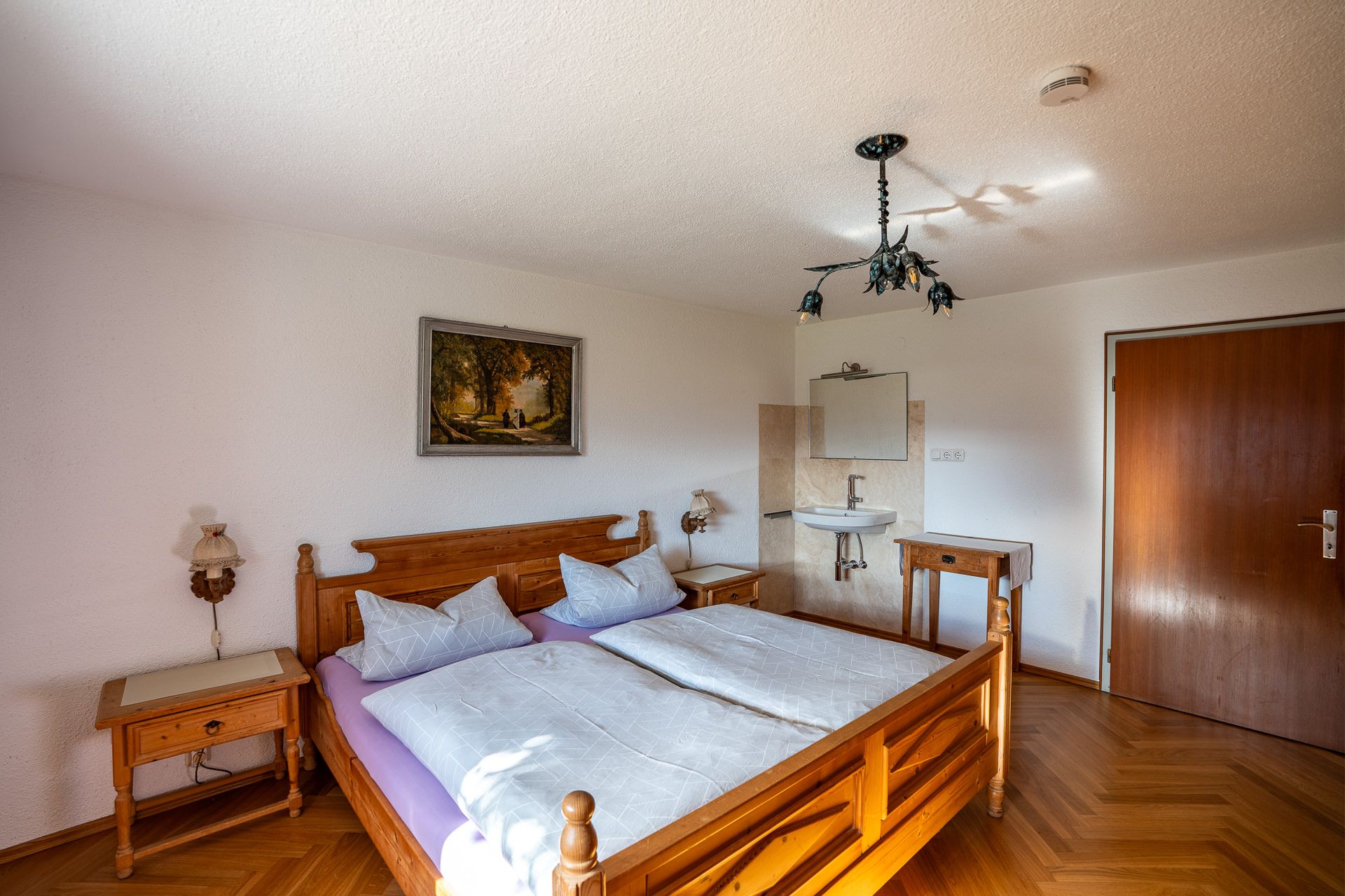 Dasselbe Schlafzimmer mit dem Doppelbett aus hellem Holz, hinten Waschbecken und Spiegel, ein Tischchen und die Zimmertür