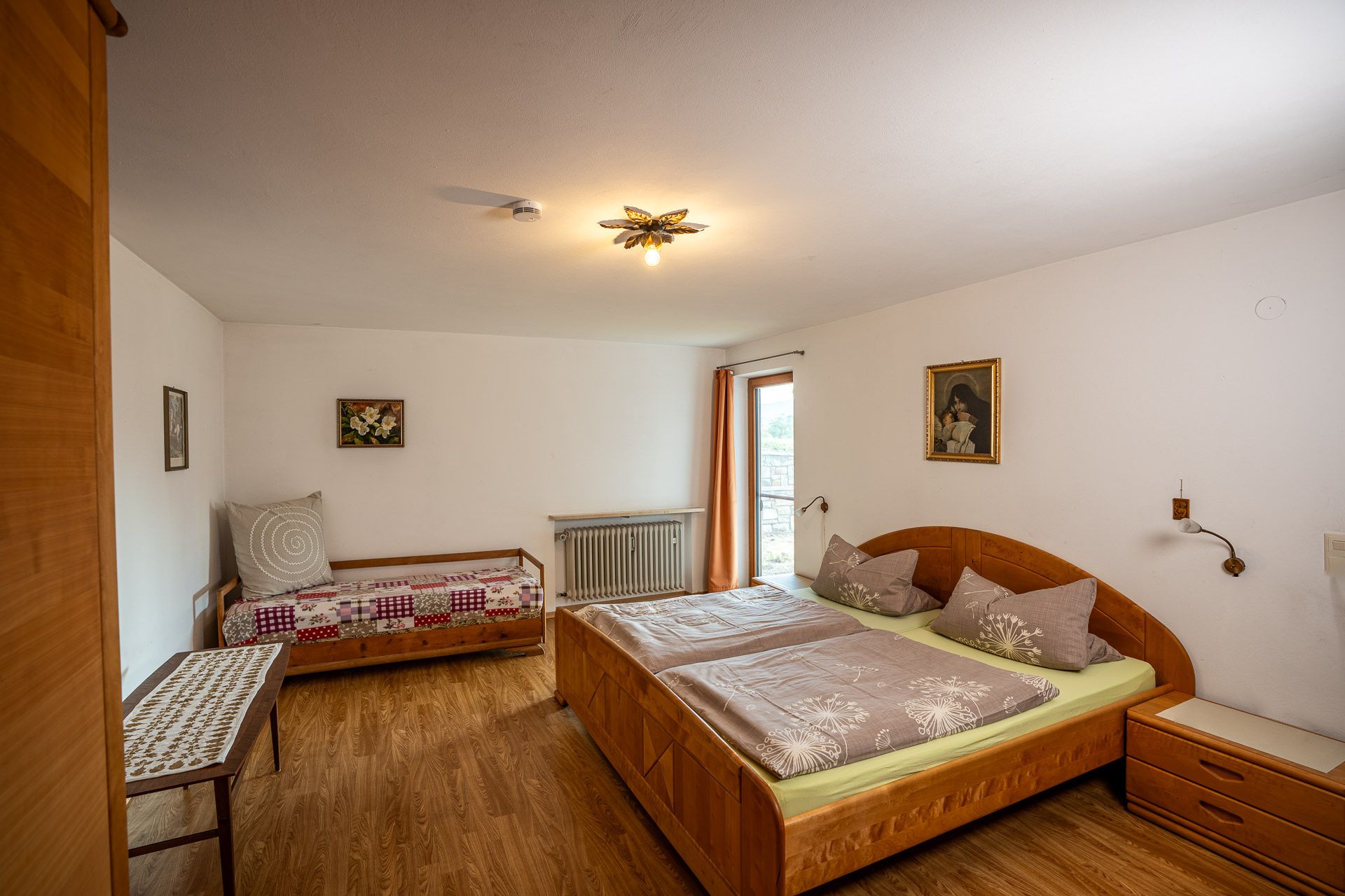 Zweites Schlafzimmer mit Doppelbett aus Holz und einem Einzelbett mit bunter Patchworkdecke, hinten ein bodentiefes Fenster mit Vorhang