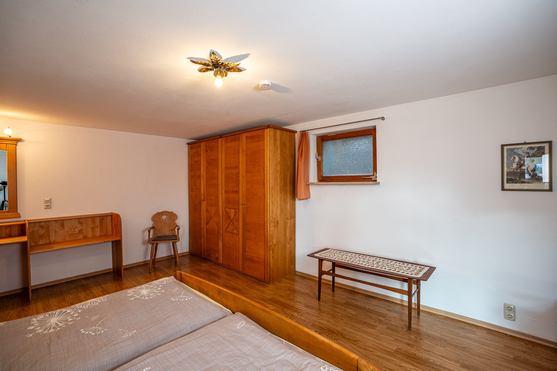 Hoher Kleiderschrank aus Holz im zweiten Schlafzimmer, daneben ein kleines Fenster mit Milchglas, ein niedriger Tisch mit Läufer und ein Bild an der Wand