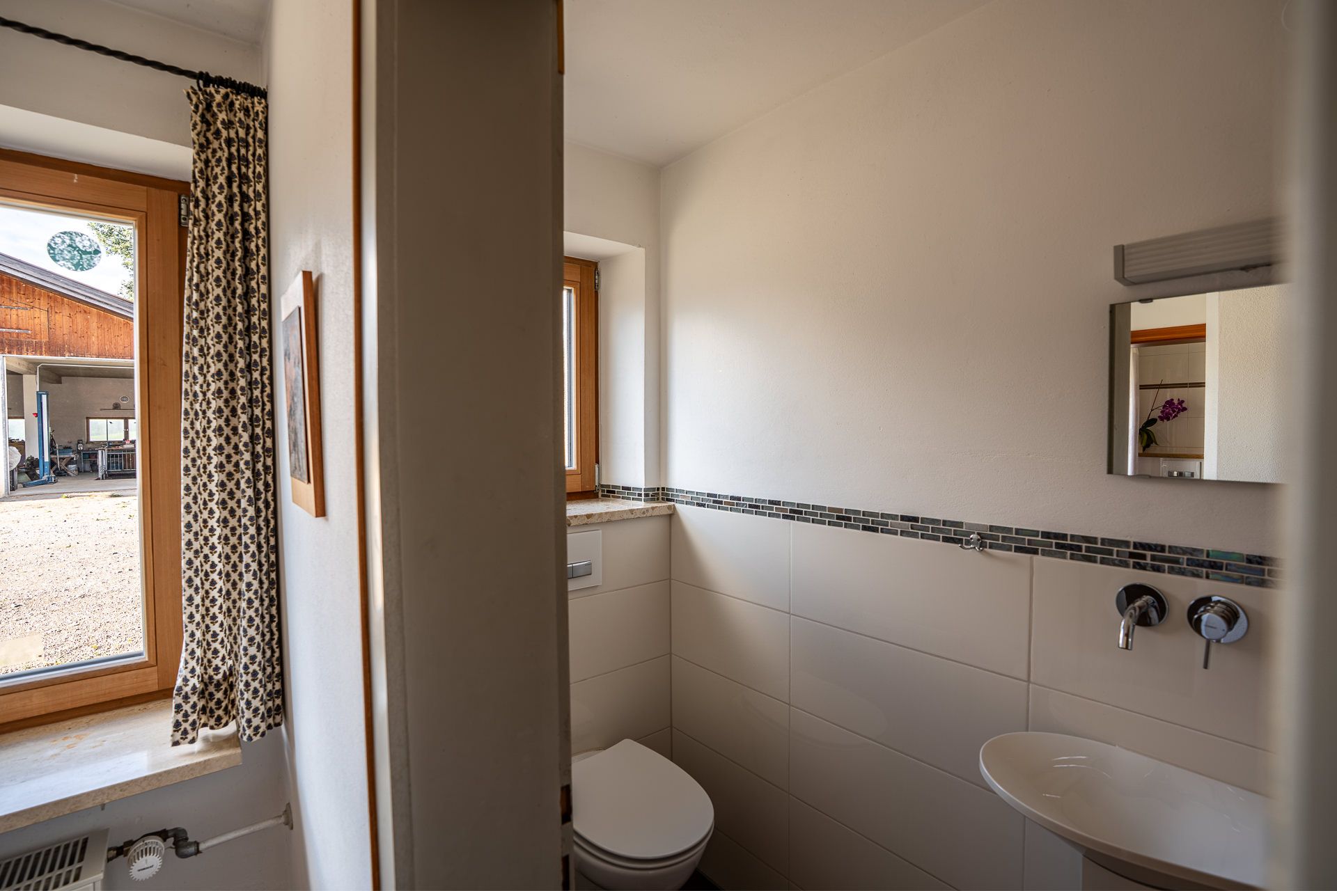 Separates WC mit kleinem Waschbecken und Spiegel, links ein Fenster mit Vorhang