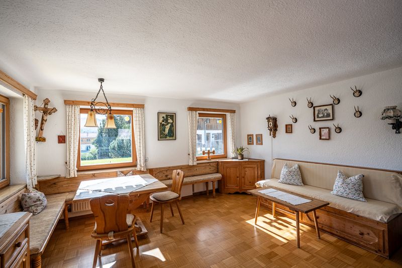 Wohnzimmer mit Eckbank und Tisch unter dem Kruzifix im Herrgottswinkel, rechts ein langes Sofa mit Kissen, darüber Geweihe, eine Wanduhr mit Gewichten und alte Fotos, dazwischen zwei Fenster