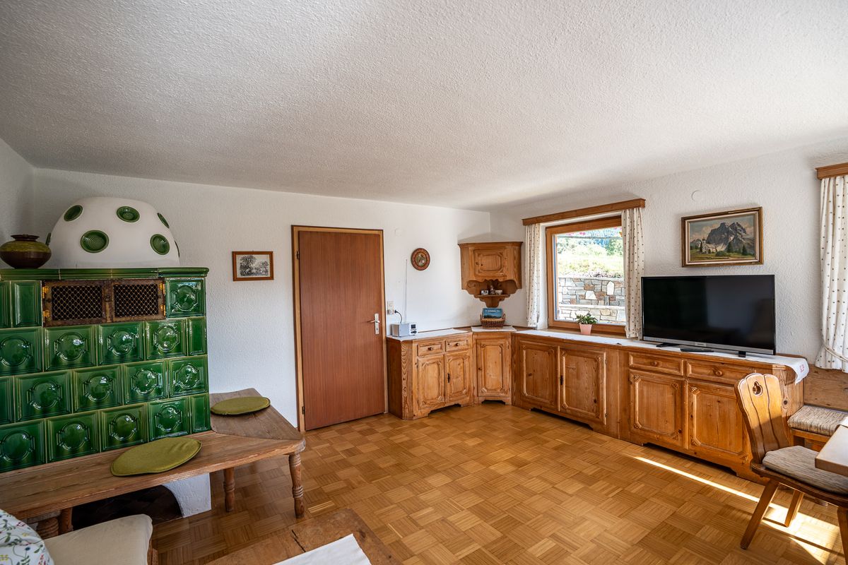 Der grüne Kachelofen mit Ofenbank und Sitzkissen, daneben Holzschränke unter dem Fenster, ein Fernseher und ein Bergbild an der Wand