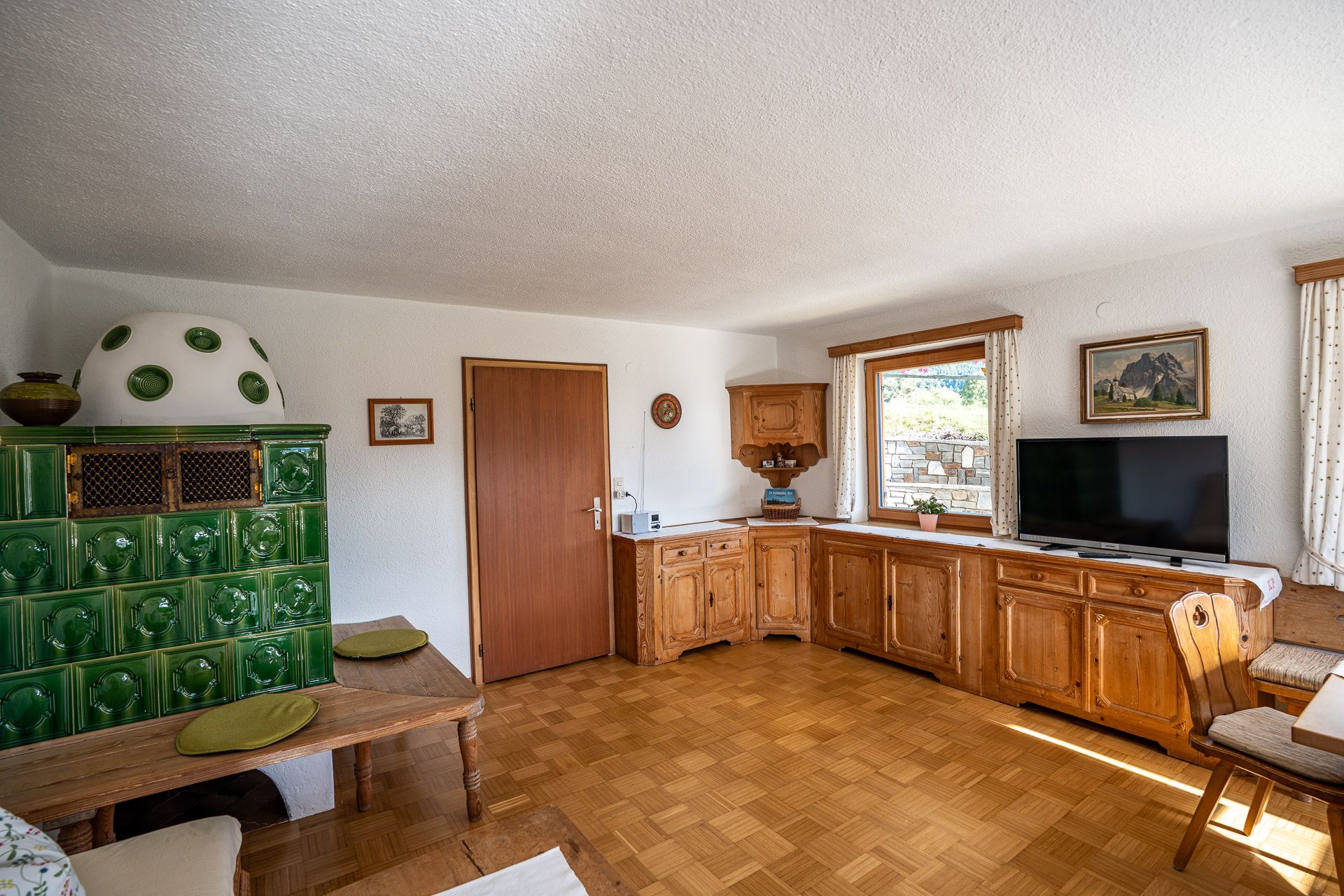Der grüne Kachelofen mit Ofenbank und Sitzkissen, daneben Holzschränke unter dem Fenster, ein Fernseher und ein Bergbild an der Wand