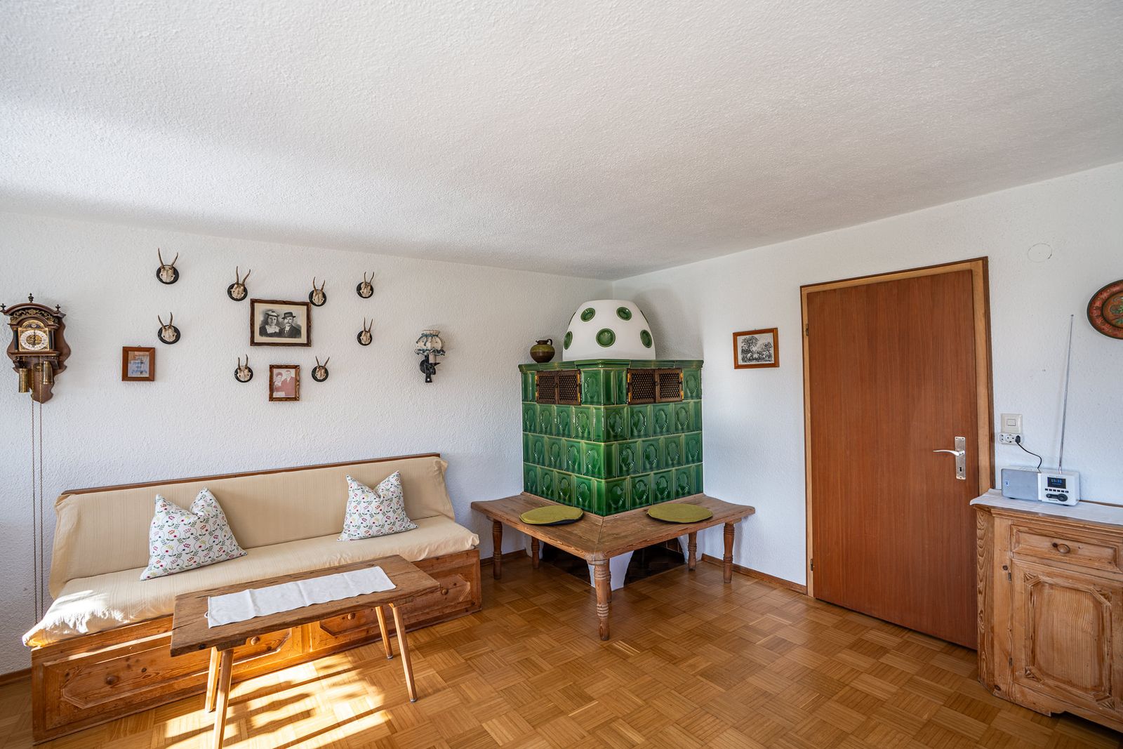 Living room of the ground-floor apartment with the green tiled stove and its bench in the corner, a sofa with cushions on the left and a weight-driven clock and antlers on the wall