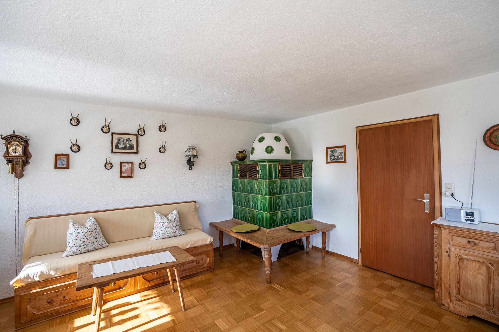 Wohnzimmer der Ferienwohnung im Erdgeschoss mit dem grünen Kachelofen und der Ofenbank in der Ecke, links ein Sofa mit Kissen und ein Holztisch, an der Wand eine Uhr mit Gewichten, Geweihe und alte Fotos