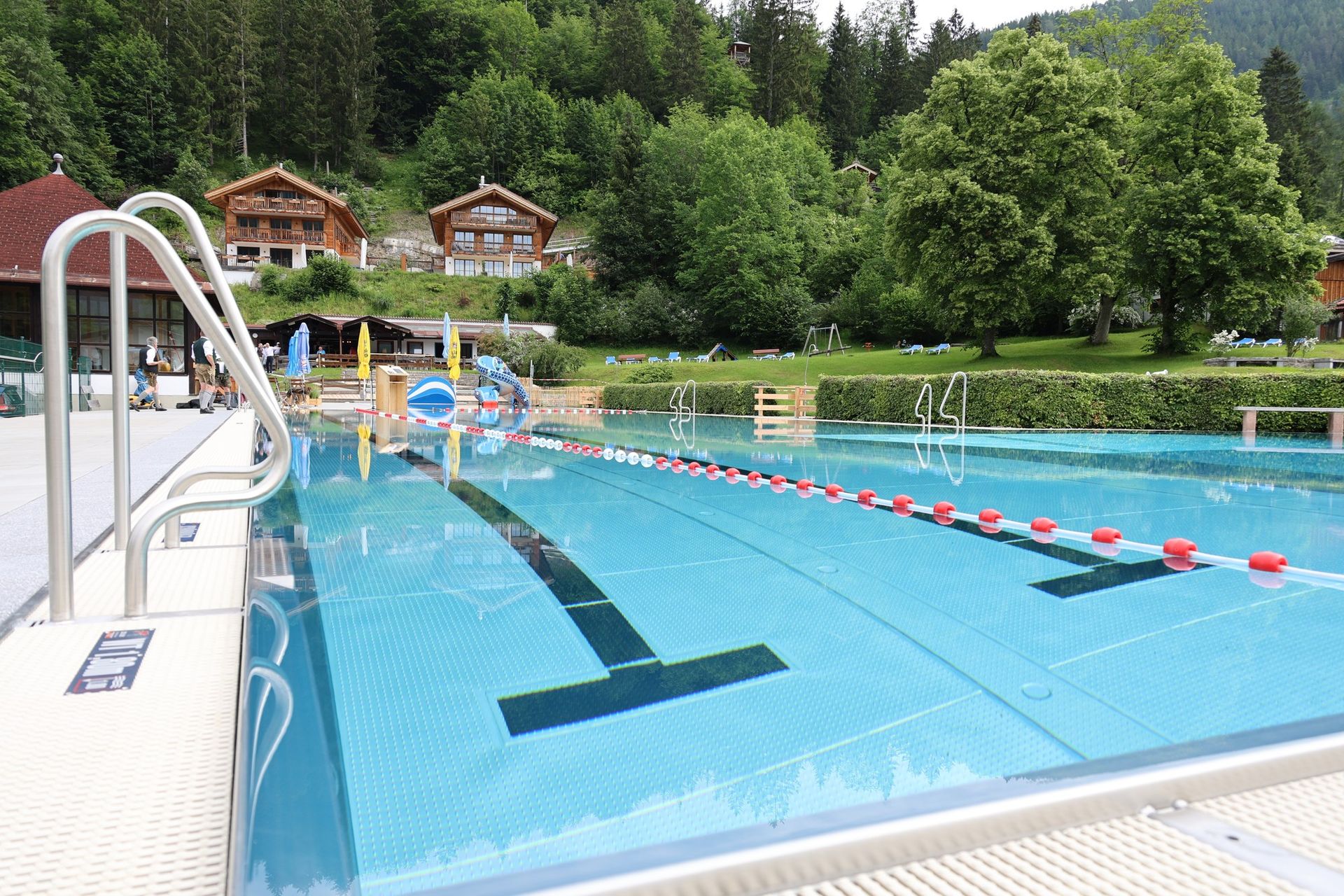 Schwimmerbecken des Alpenfreibads Bayrischzell mit Leinen und Einstiegsleiter, dahinter Rutsche, Liegewiese und Holzhäuser am Waldhang