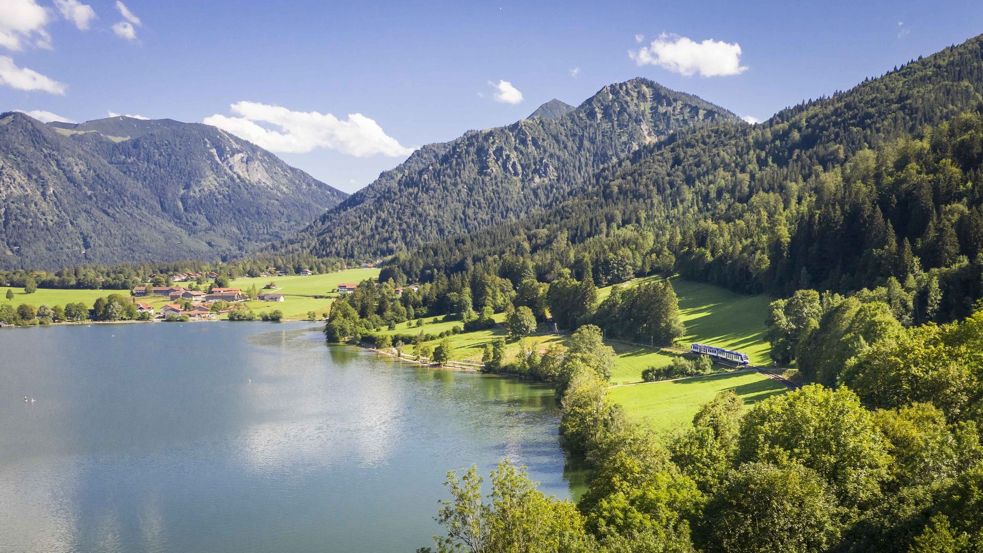 Luftbild vom Südufer des Schliersees mit Wiesen und Wald, rechts fährt klein ein blau-weißer Zug der BRB, dahinter bewaldete Berge