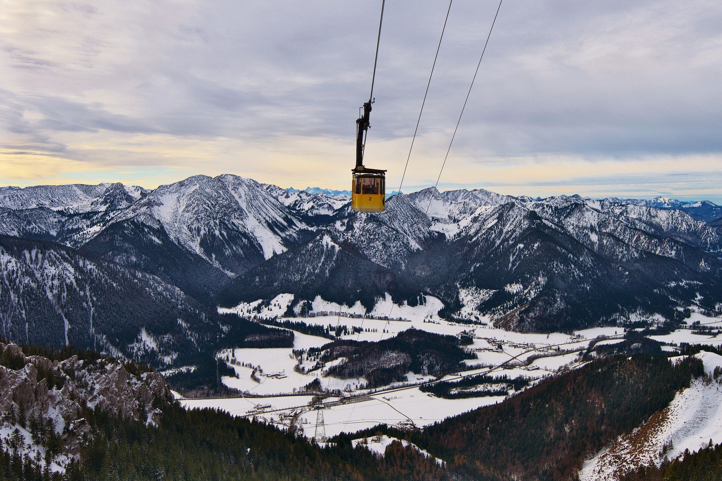 Gelbe Gondel am Seil vor verschneiten Bergketten, darunter das winterliche Tal mit Wiesen und Wald im Abendlicht