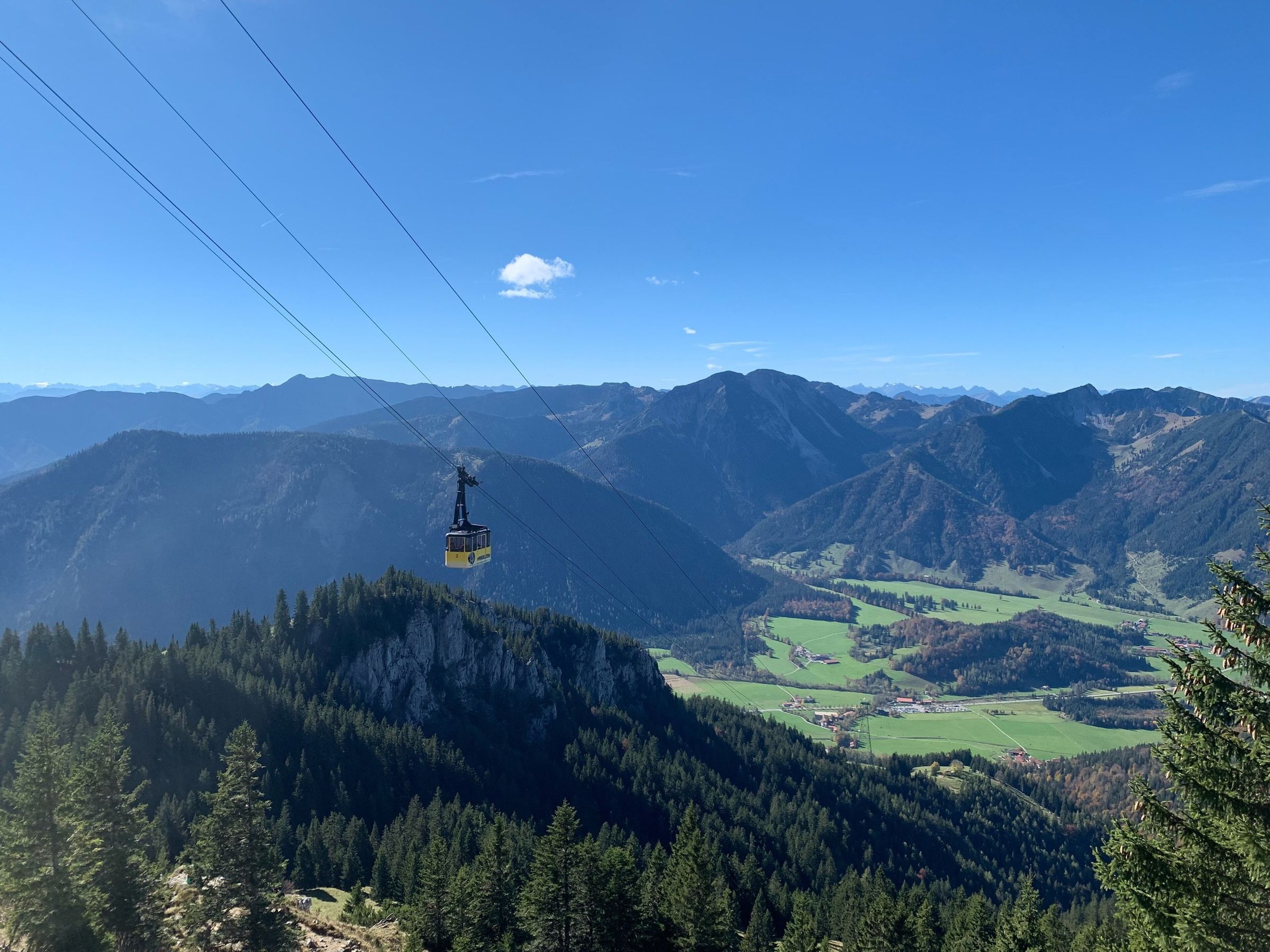 Gelbe Gondel der Seilbahn über Bergwald und einem Felskopf, unten die grünen Wiesen von Osterhofen, dahinter Bergketten unter blauem Himmel