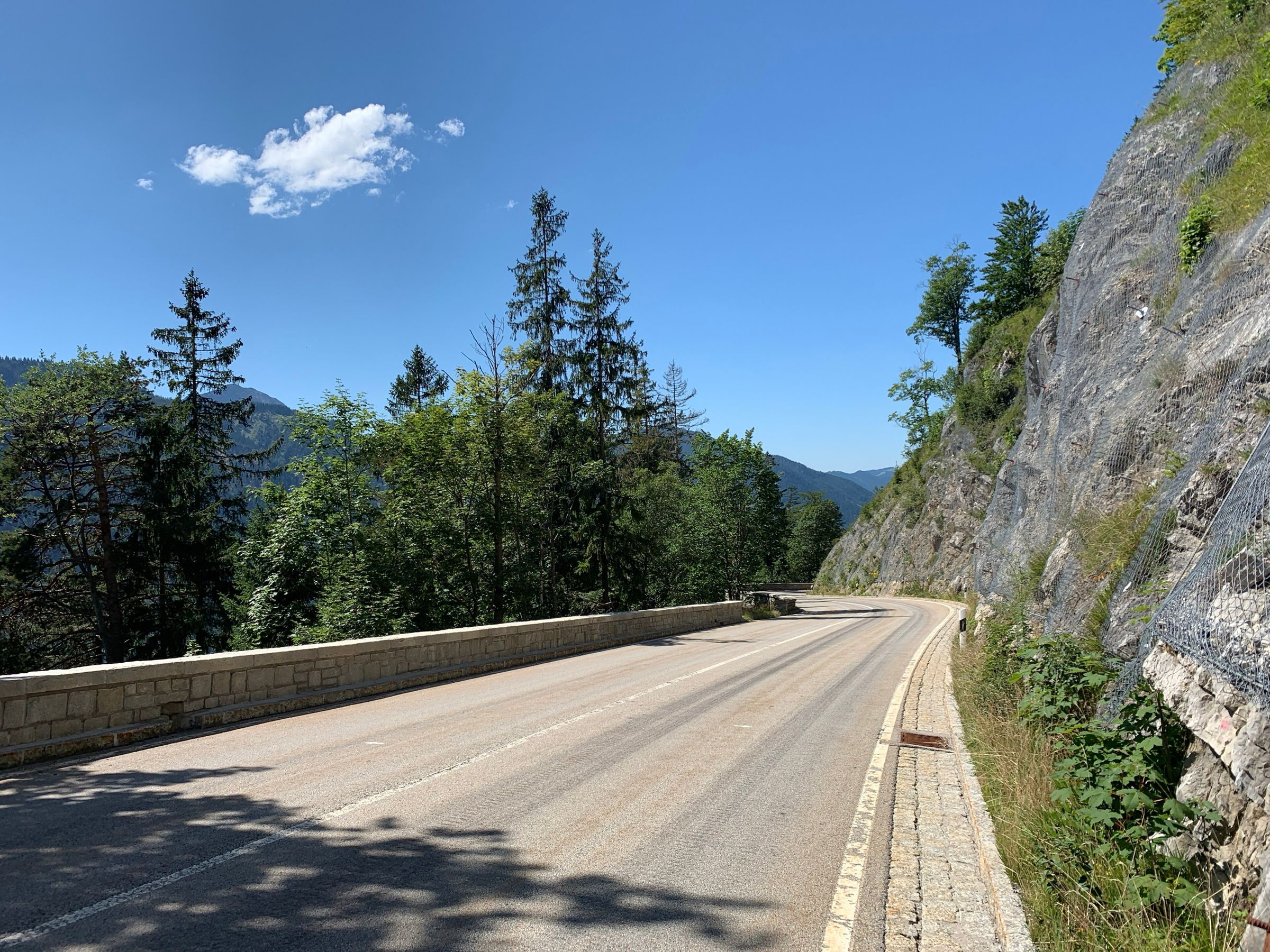 Leere Passstraße mit Steinmauer entlang einer Felswand, Bergwald und tiefblauer Himmel