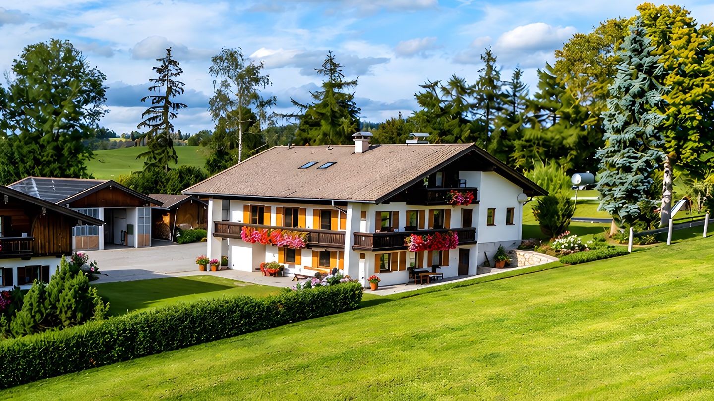 Aerial view of the holiday house with wooden balconies and geraniums, the paved farm drive between the farm building and the holiday house and an outbuilding with open doors on the left, a hedge and the meadow in front, trees behind