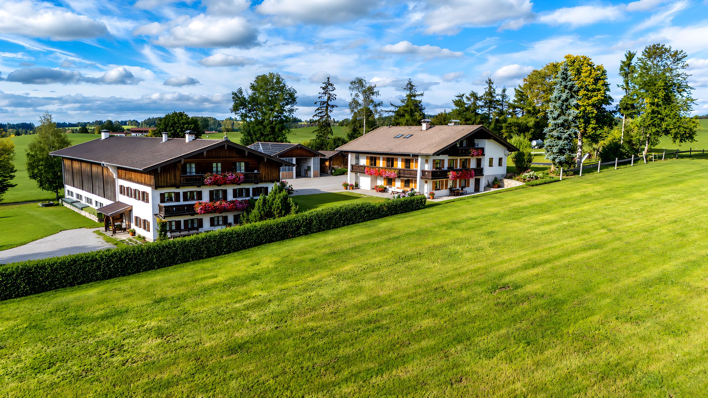 Luftbild: Stallgebäude und Ferienhaus mit Geranien an den Balkonen, davor eine Hecke und eine große Wiese, dahinter Bäume und ein Himmel mit Wolken