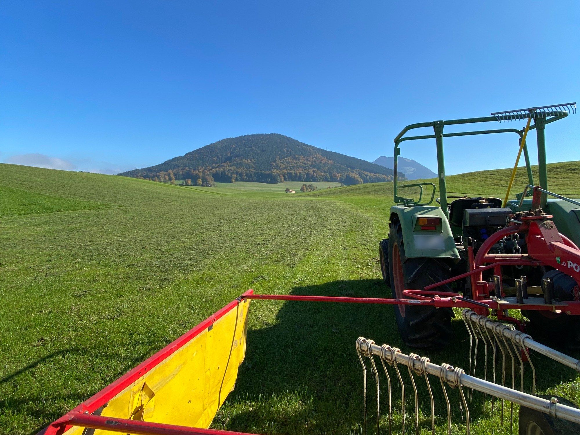 Ein älterer grüner Traktor mit rotem Heuwender auf der gemähten Wiese, dahinter ein bewaldeter Berg unter blauem Himmel