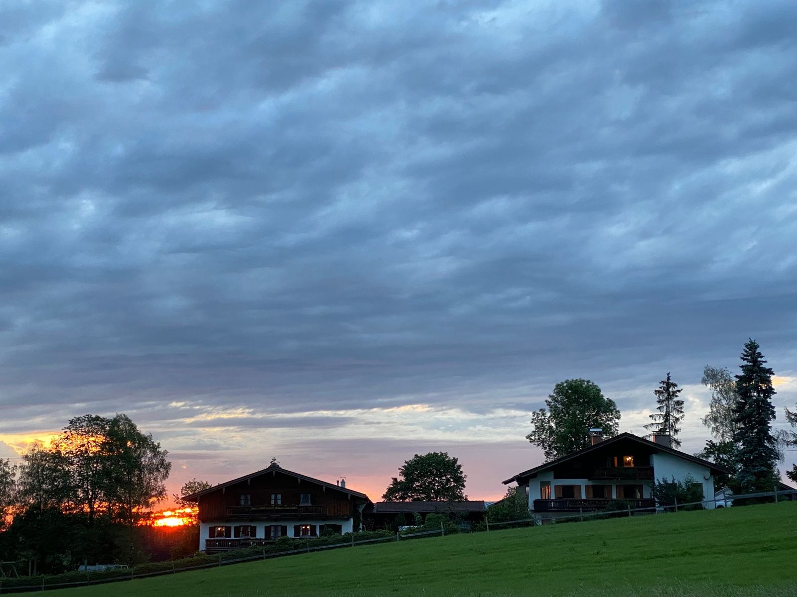 Bauernhaus und Ferienhaus in der Abenddämmerung unter grauen Wolken, links am Horizont leuchtet das Abendrot