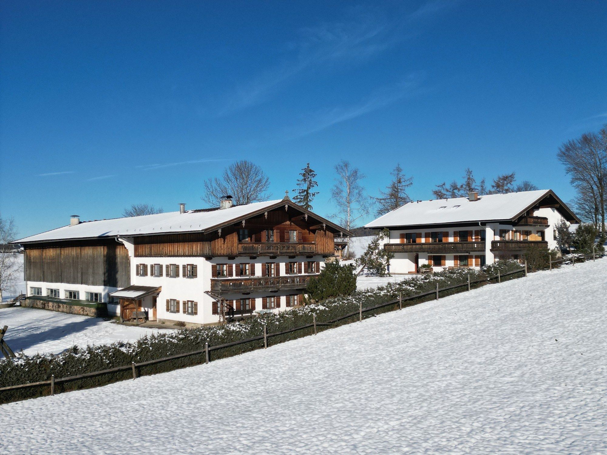 Der Hof im Schnee aus der Luft: Bauernhaus und Ferienhaus mit verschneiten Dächern unter tiefblauem Himmel, davor ein verschneiter Hang