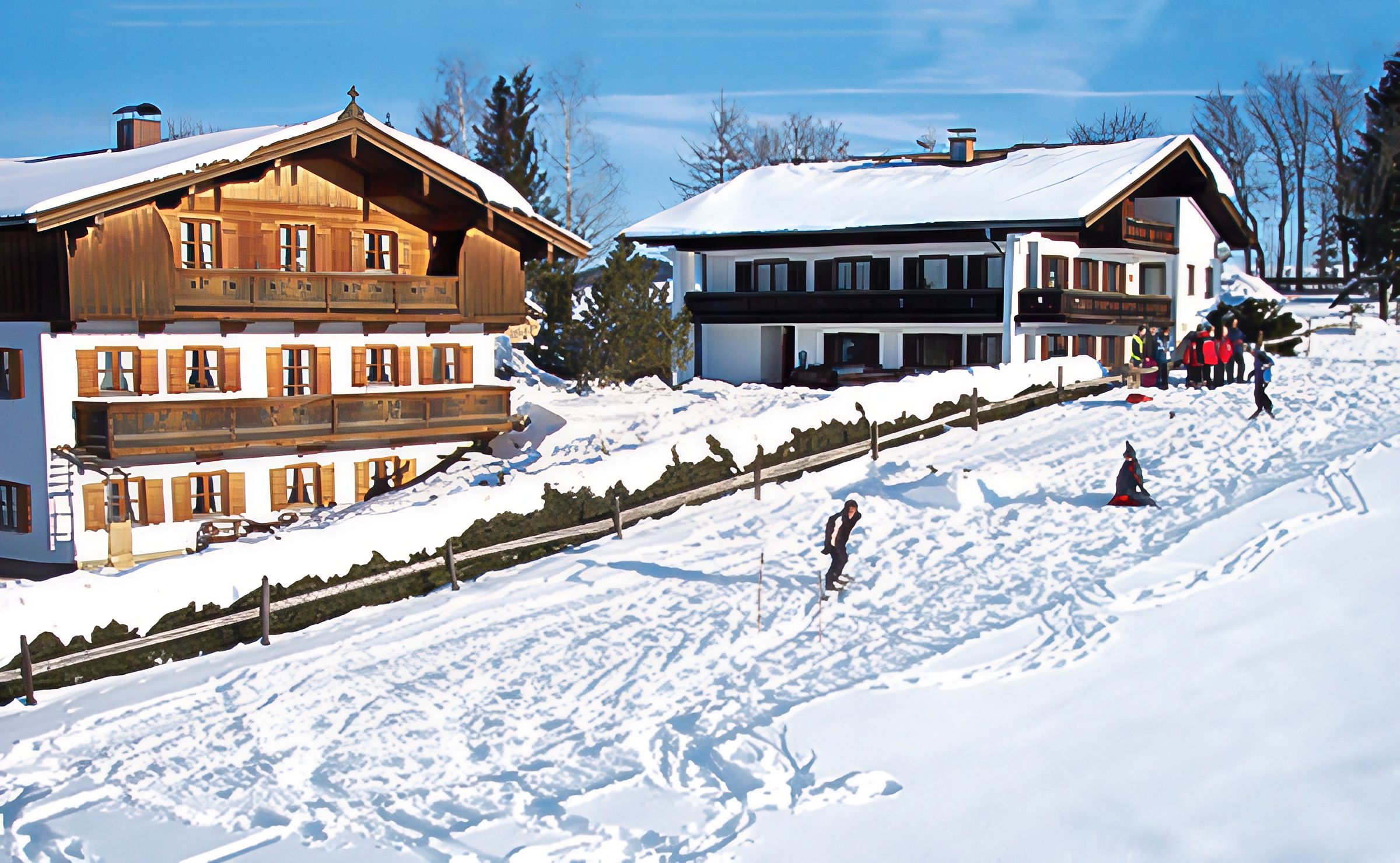 Bauernhof und Ferienhaus im Winter, davor ein verschneiter Hang, auf dem Kinder rodeln und Ski fahren