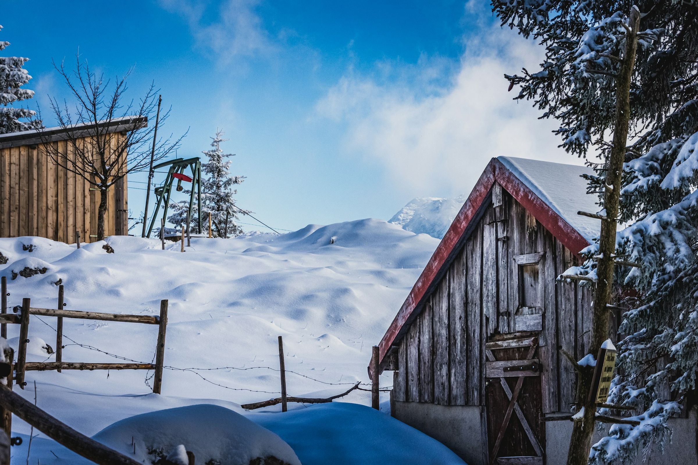 Verschneite Holzhütte mit rotem Dachrand, ein Zaun und Fichten mit Schnee unter blauem Himmel